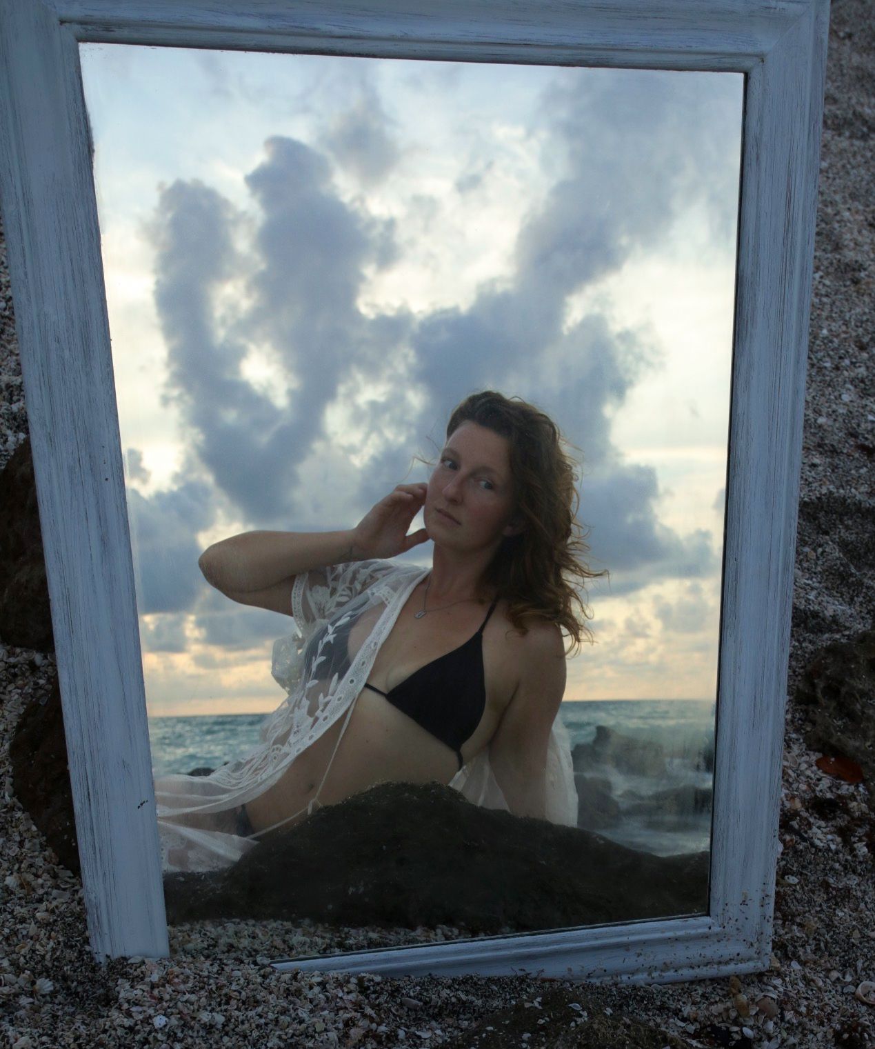 Woman in black bikini and sheer wrap poses on rocky beach, reflected in white-framed mirror under dramatic cloudy sky.