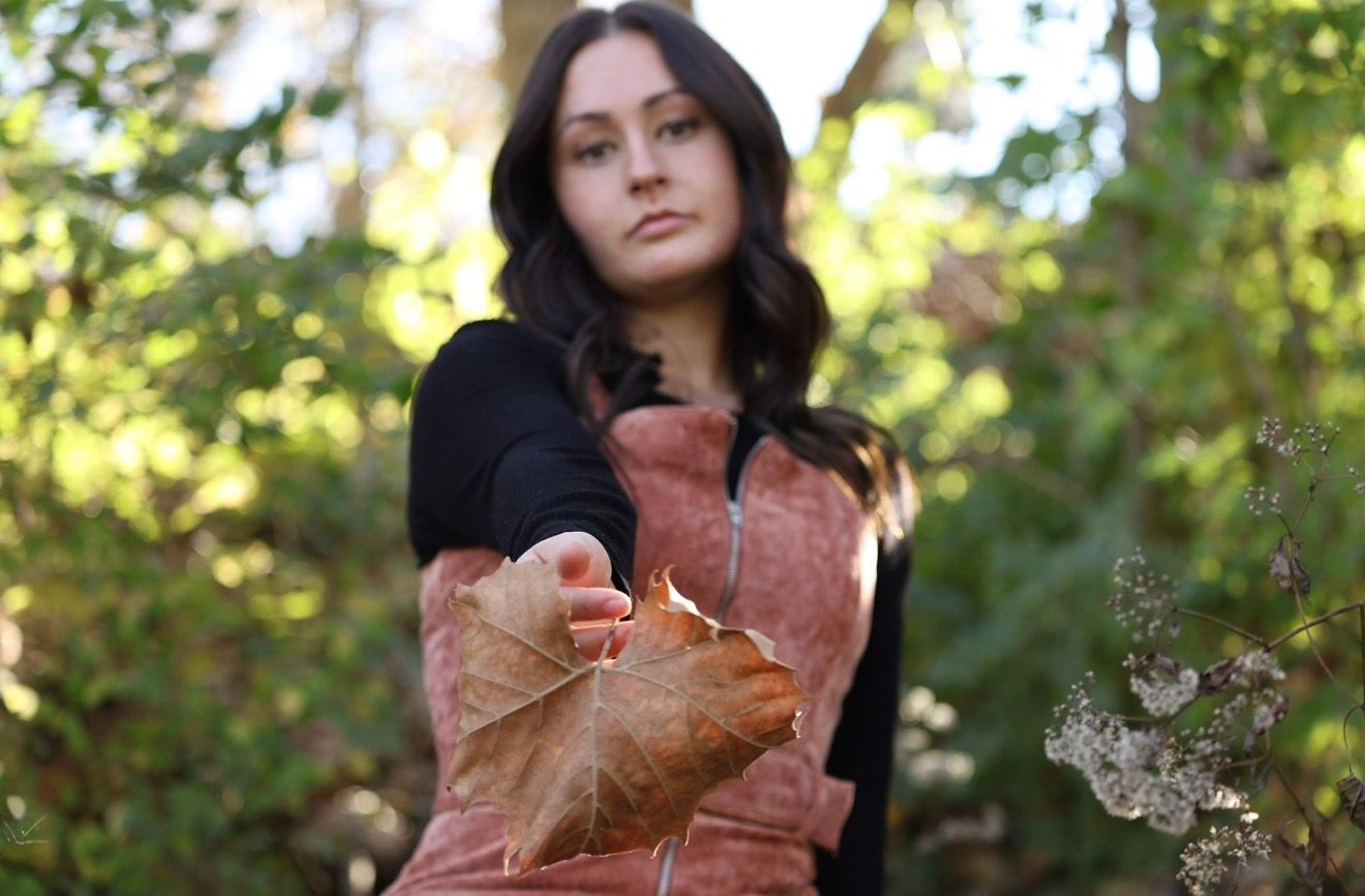 Young woman in rust vest holding a large dried autumn leaf toward camera in a sunlit forest setting.