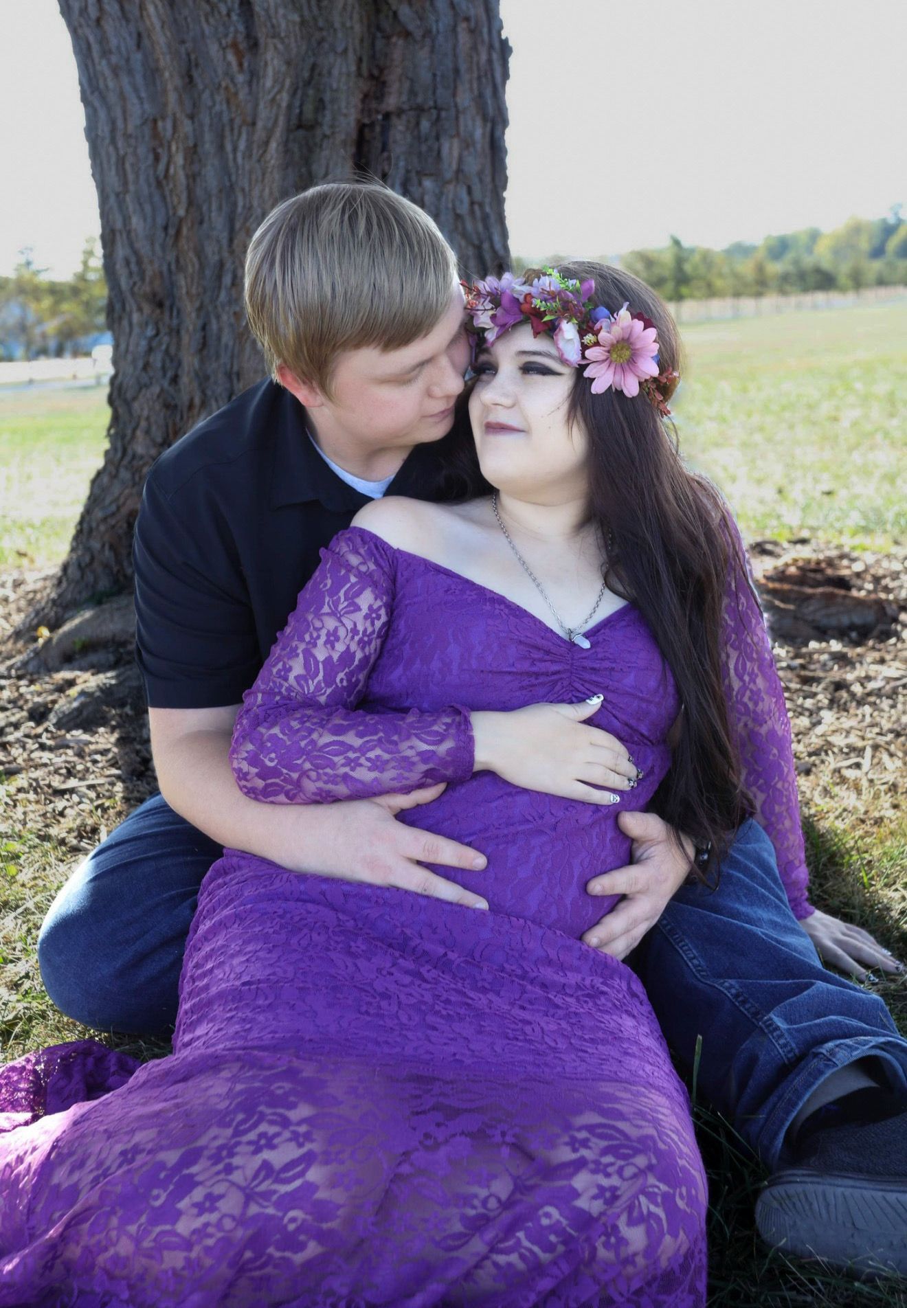 Couple sharing a tender moment outdoors, woman in purple lace maternity gown with floral crown, leaning against a tree.