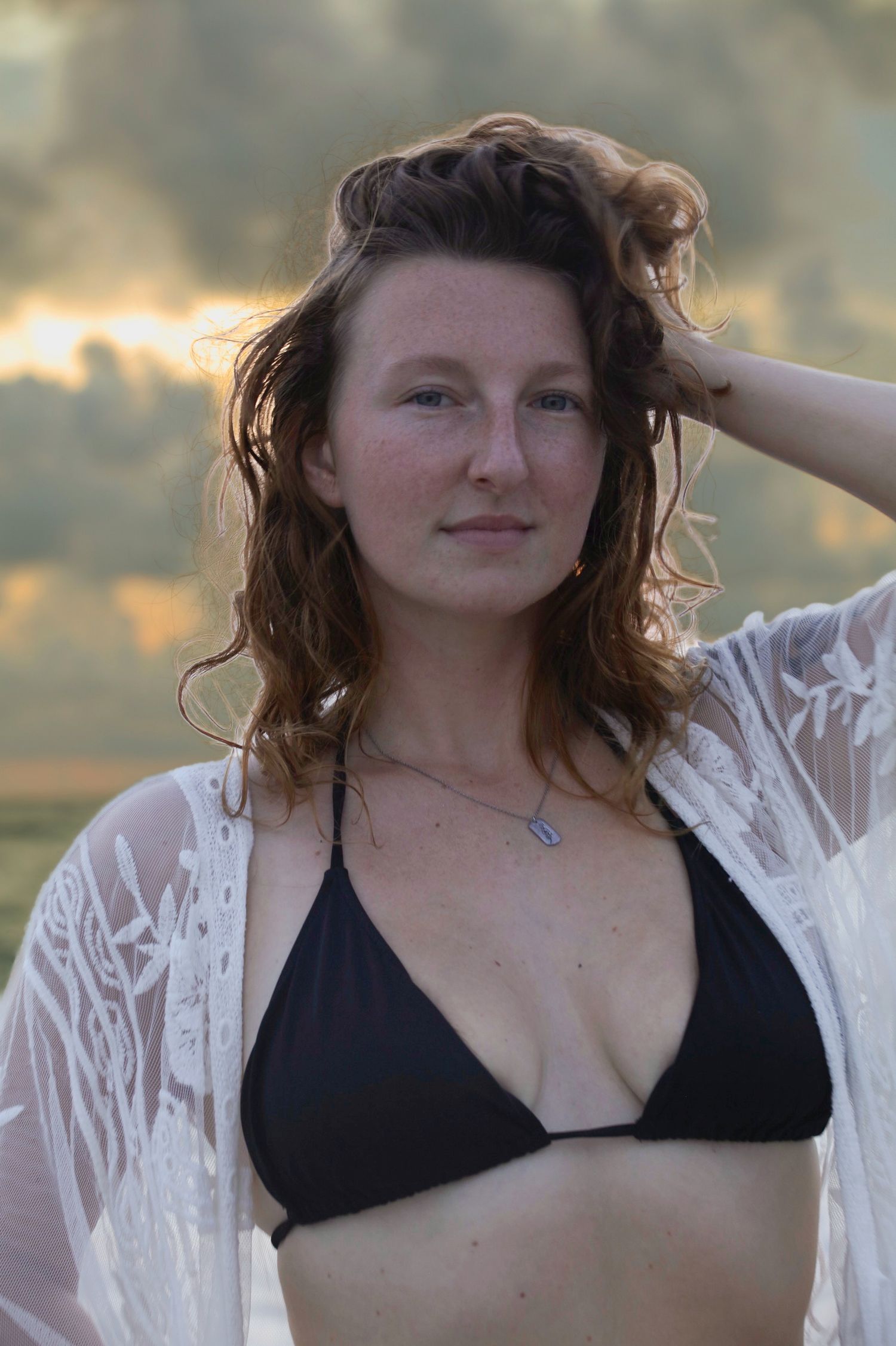 Young woman with wavy auburn hair wearing a black bikini top and white lace cover-up at the beach during sunset.