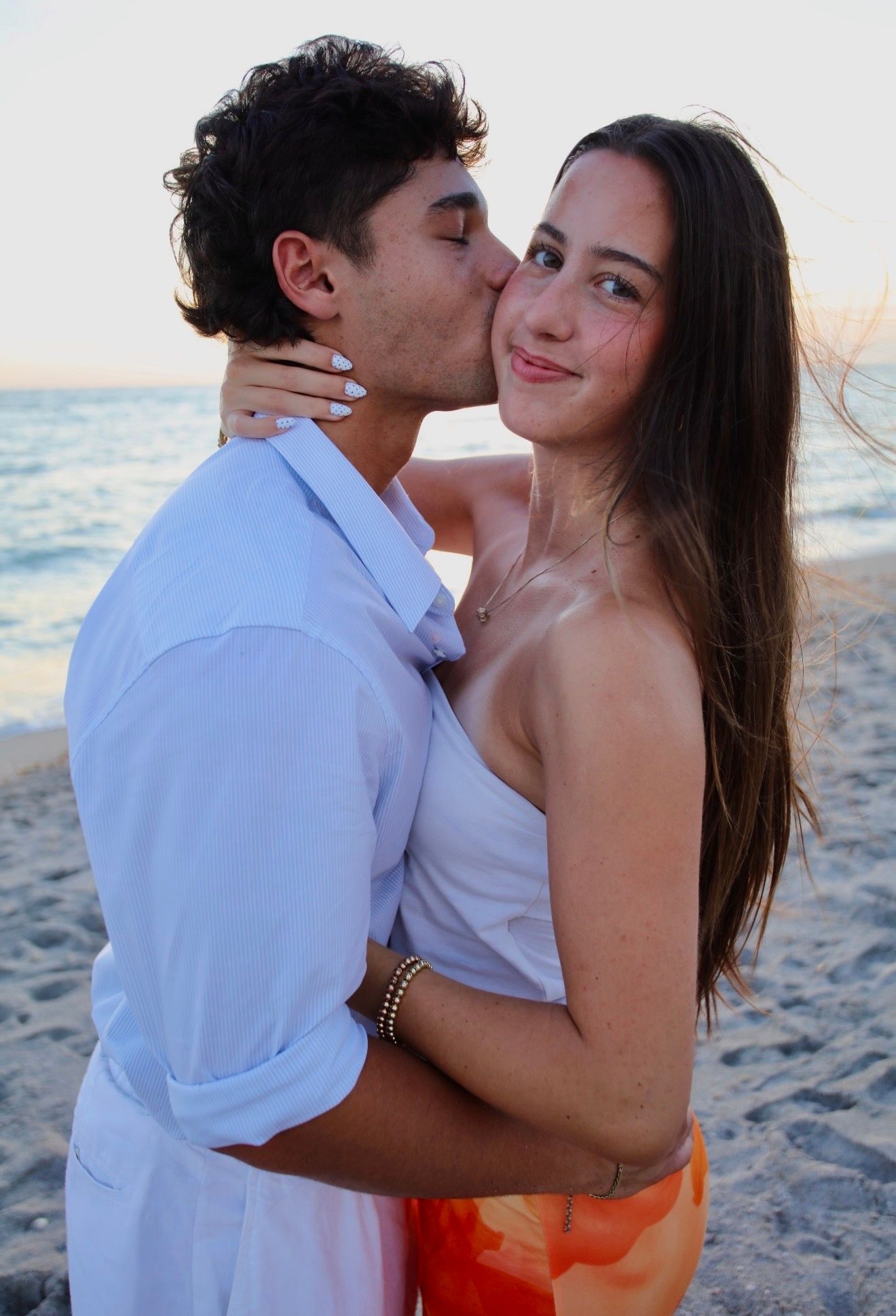 Young couple sharing a romantic kiss on the beach, woman in white top and orange shorts.