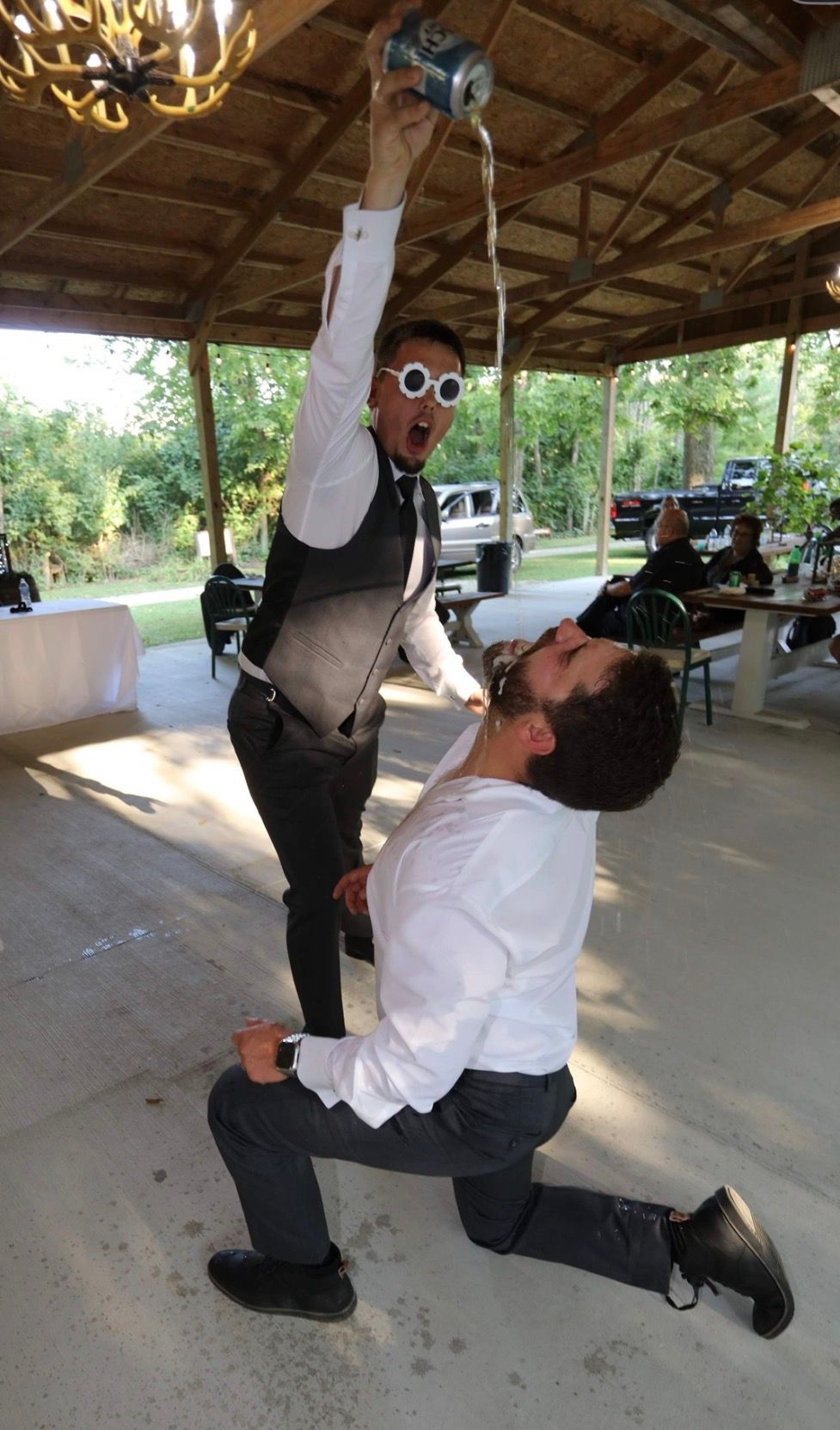 Groomsman in vest and novelty glasses pours drink into kneeling mans mouth at outdoor wedding reception pavilion.