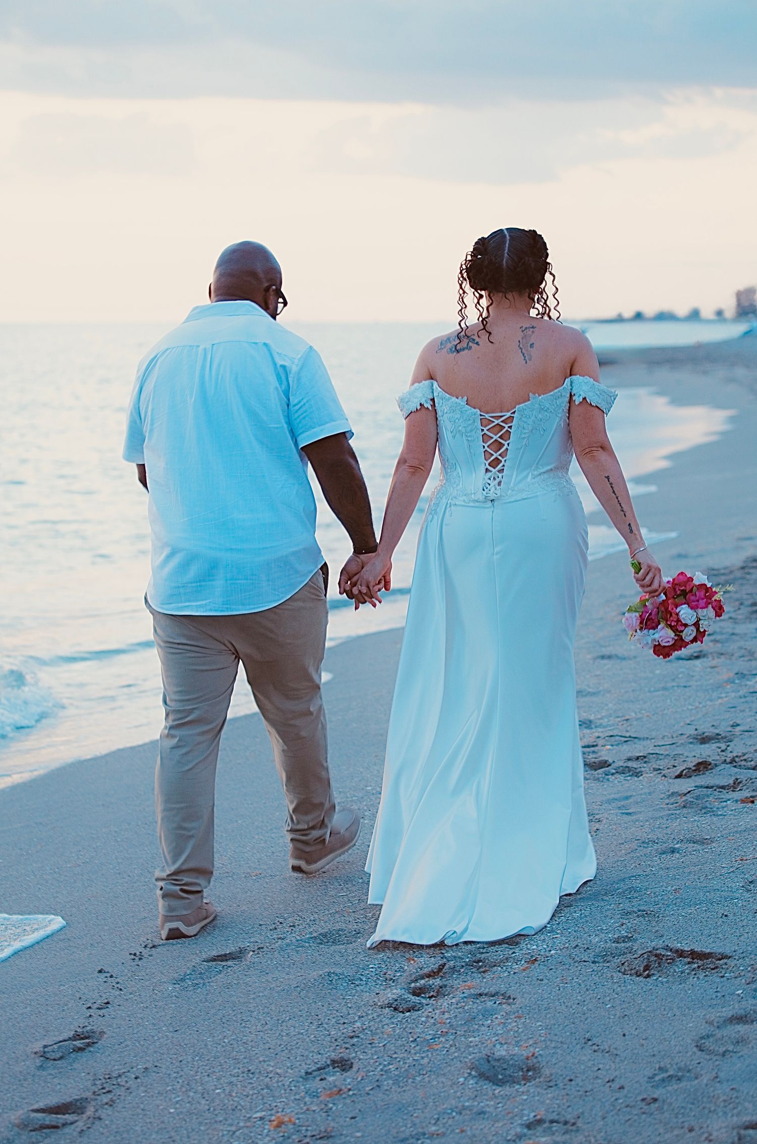 Newlywed couple holding hands walking on a beach at sunset, bride in white off-shoulder gown holding pink bouquet.