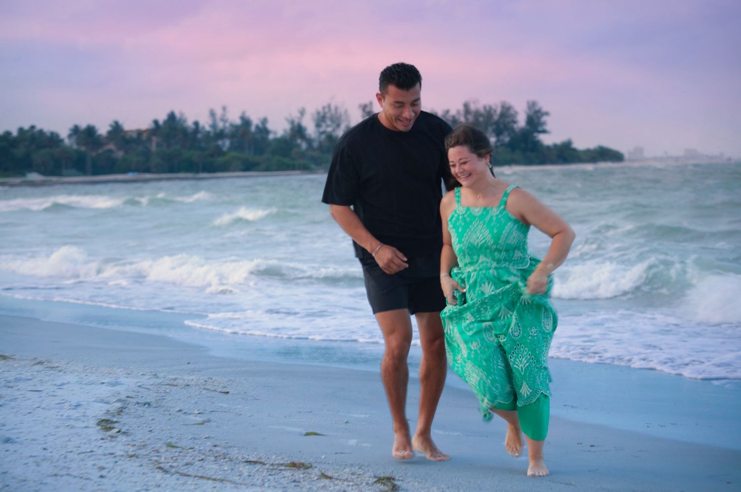 Couple walking barefoot on a sandy beach at sunset, man in black outfit and woman in green floral dress, waves behind them.