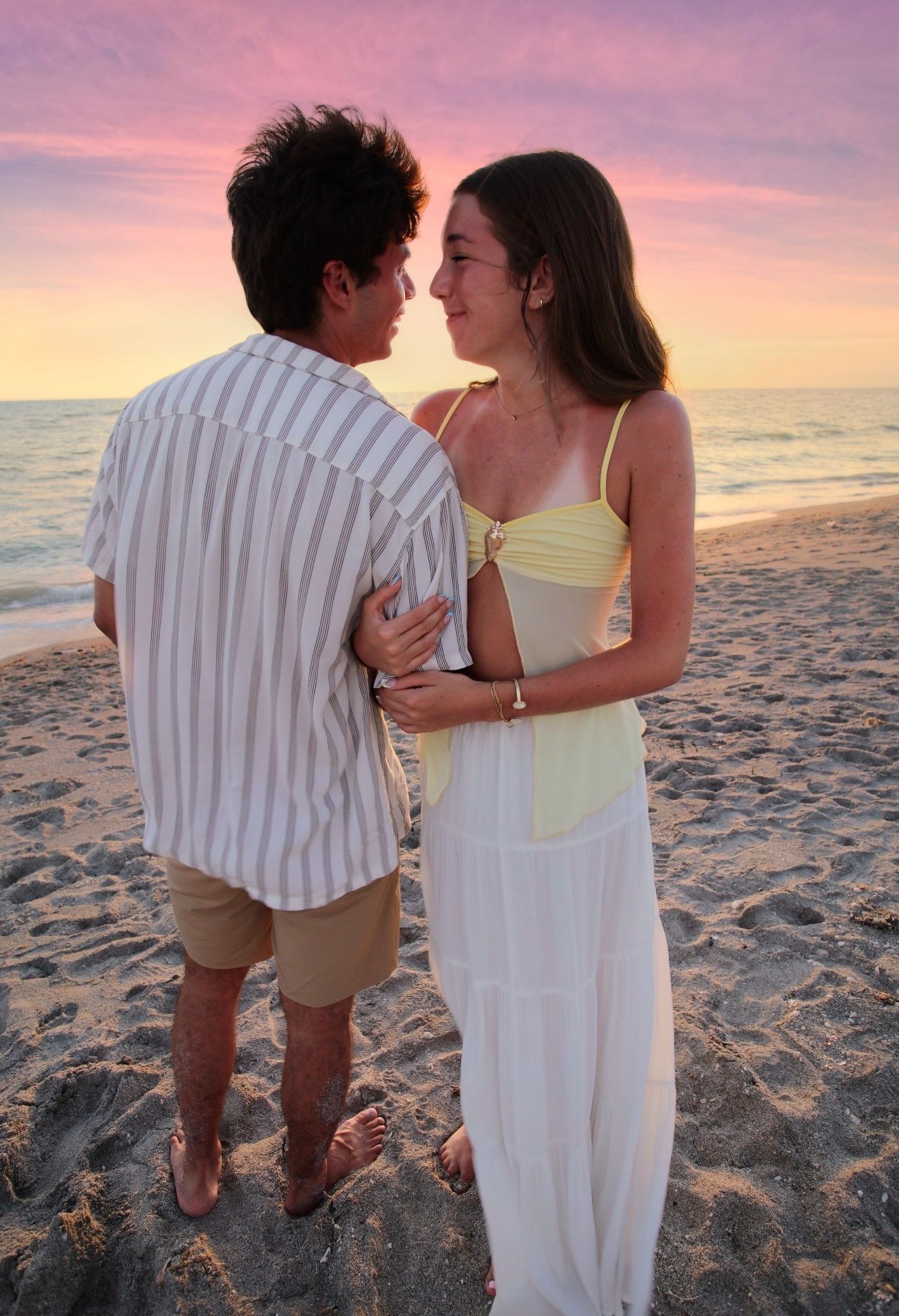 A young couple embraces and gazes at each other on a sandy beach during a colorful sunset.