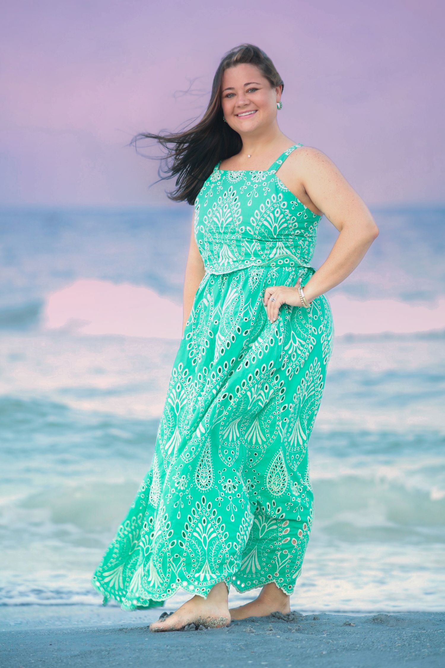 Woman in elegant green paisley maxi dress posing barefoot on beach at sunset with ocean behind her.