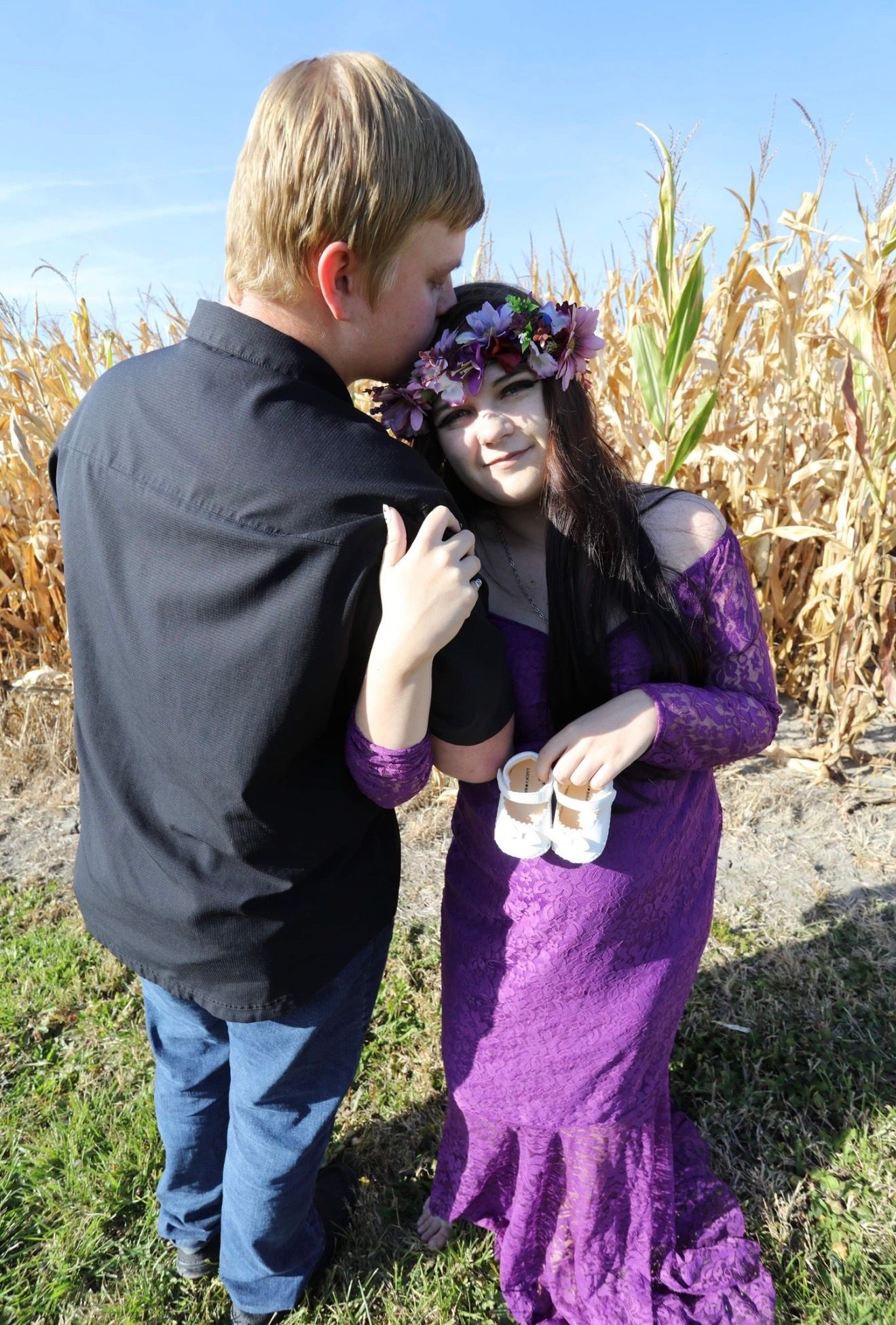 Pregnant woman in purple lace gown with floral crown shares tender moment with partner near cornfield.