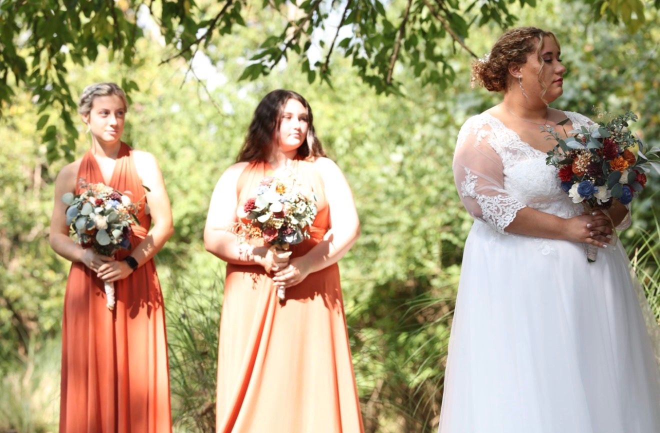 Bride in white lace gown holds bouquet with two bridesmaids in rust and peach dresses outdoors.