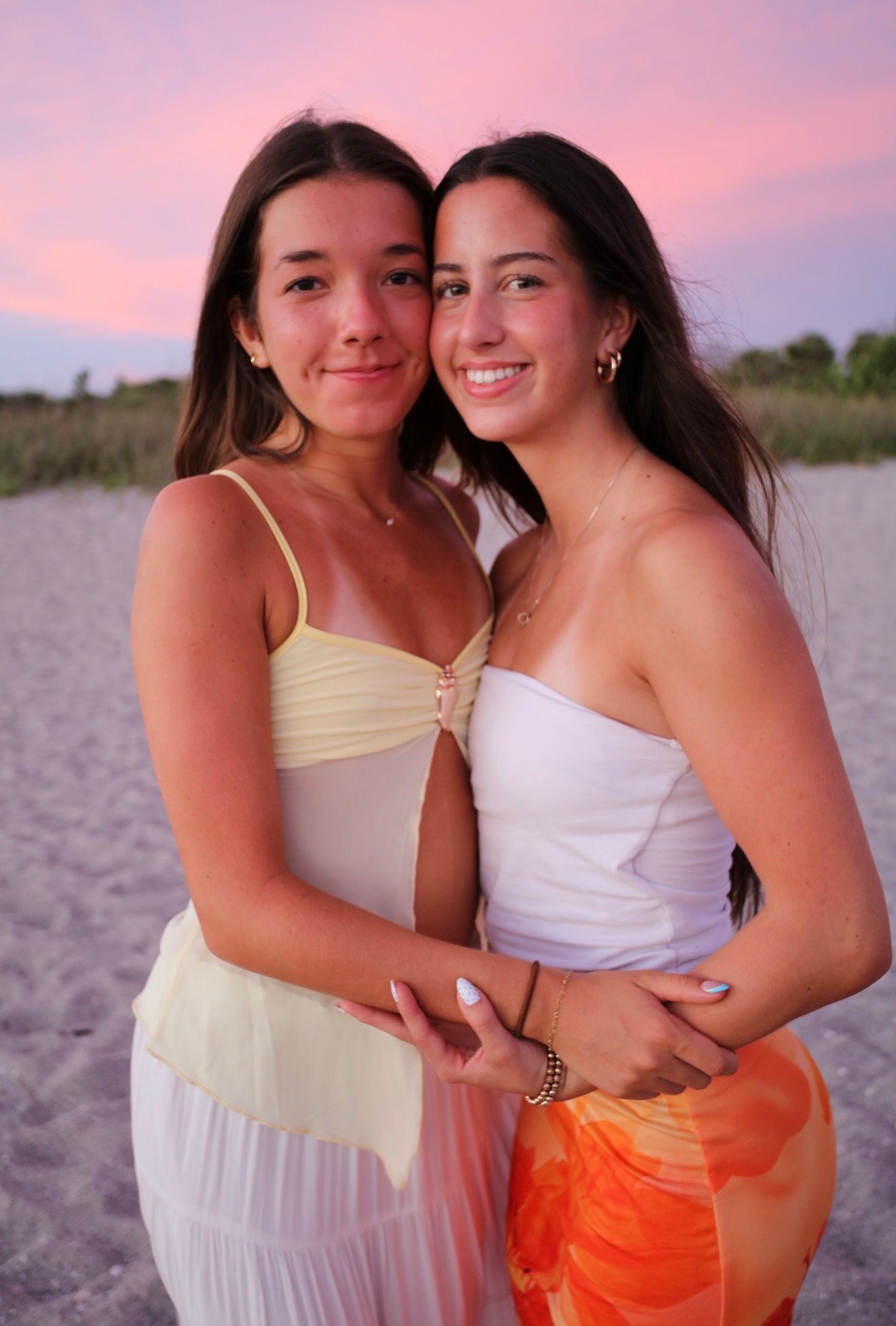 Two young women embracing on beach at sunset, one in yellow dress, one in white top and orange pants.