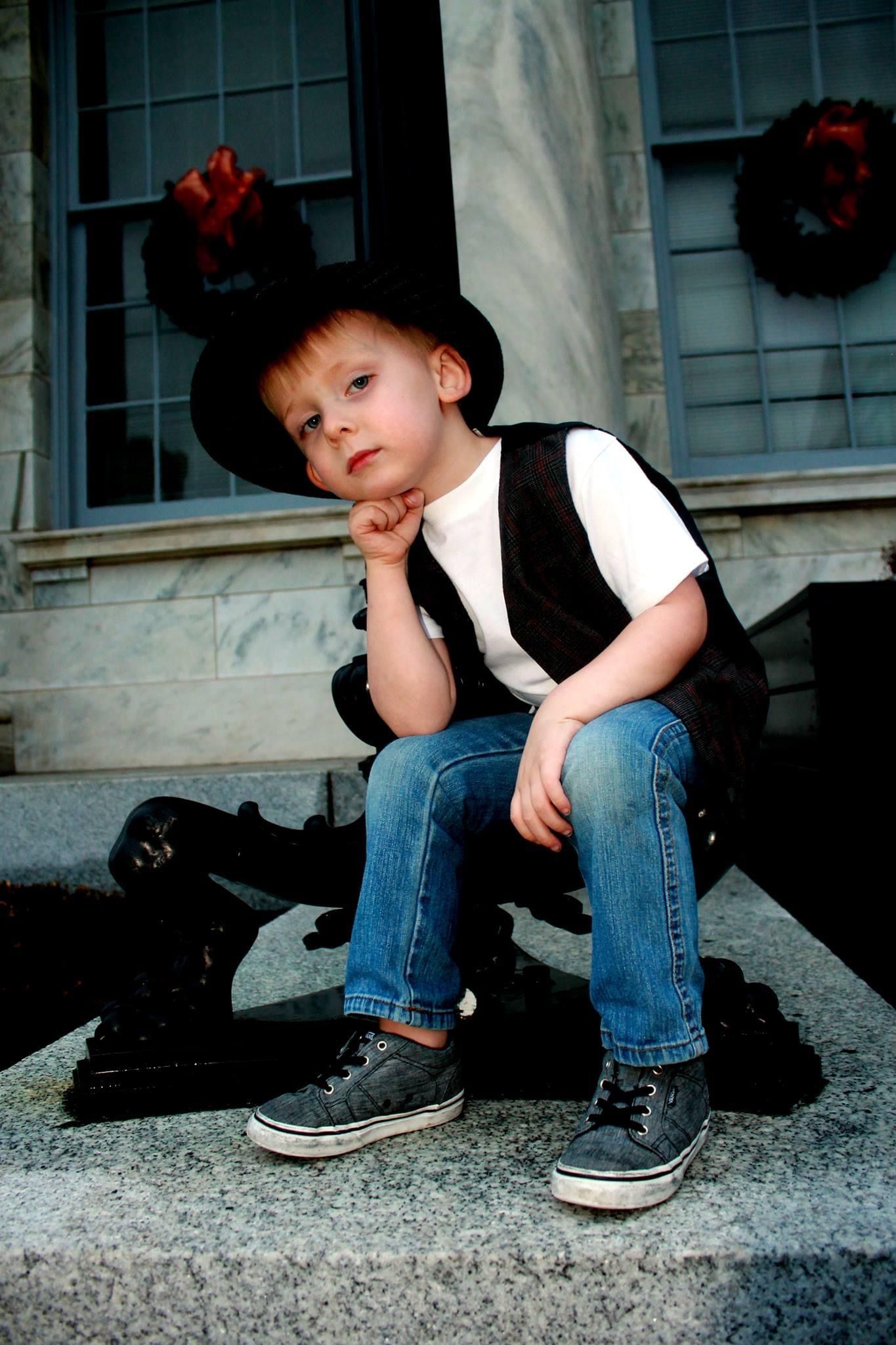 Young boy in black cowboy hat, vest, jeans, and sneakers sits thoughtfully on stone steps near holiday wreaths.