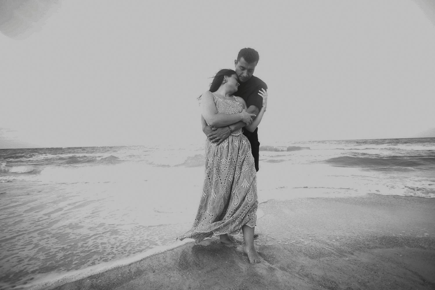Couple embracing romantically on beach shore with ocean waves in black and white photo.