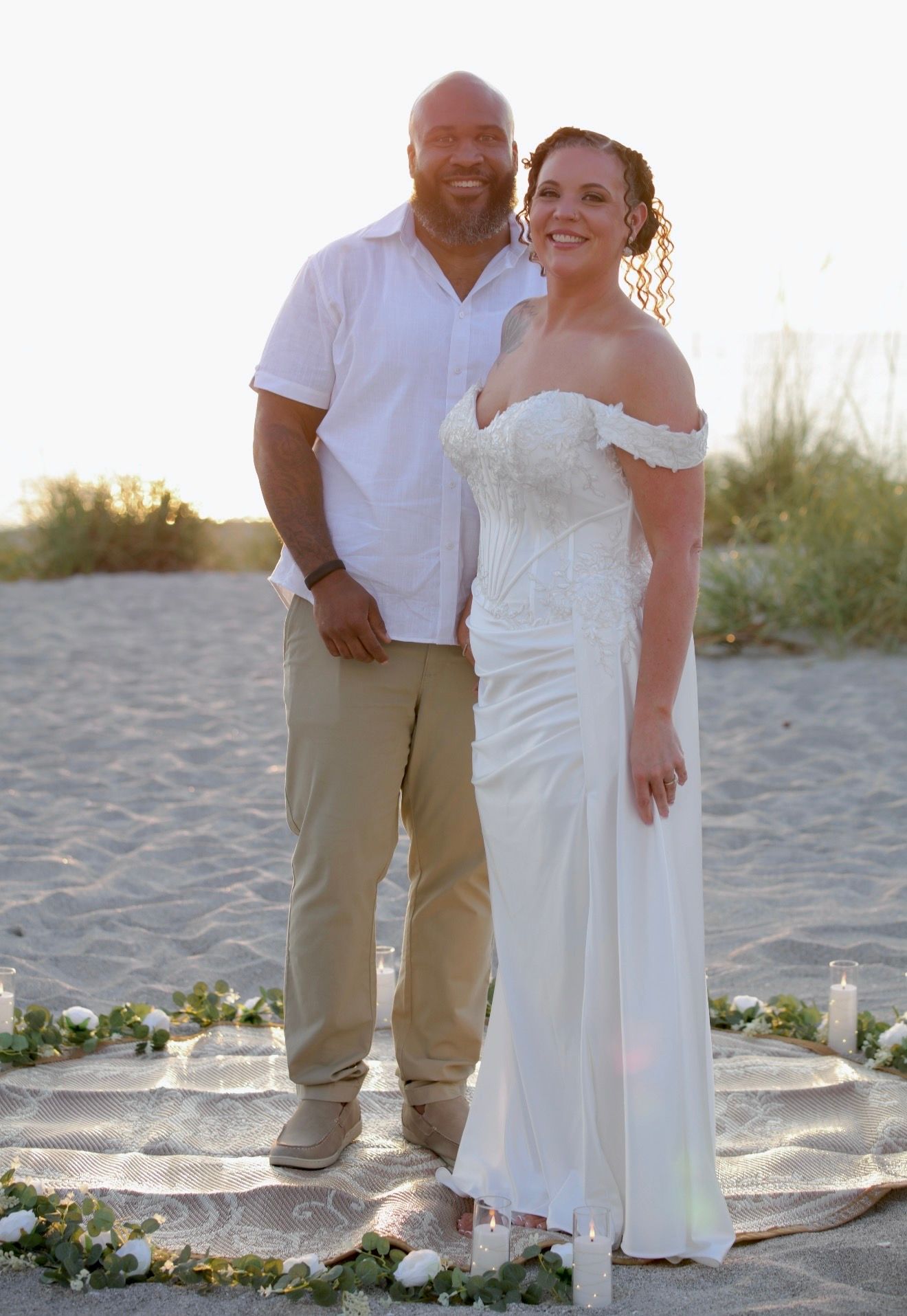 Couple posing on beach at sunset, bride in off-shoulder white gown and groom in white shirt, khaki pants.