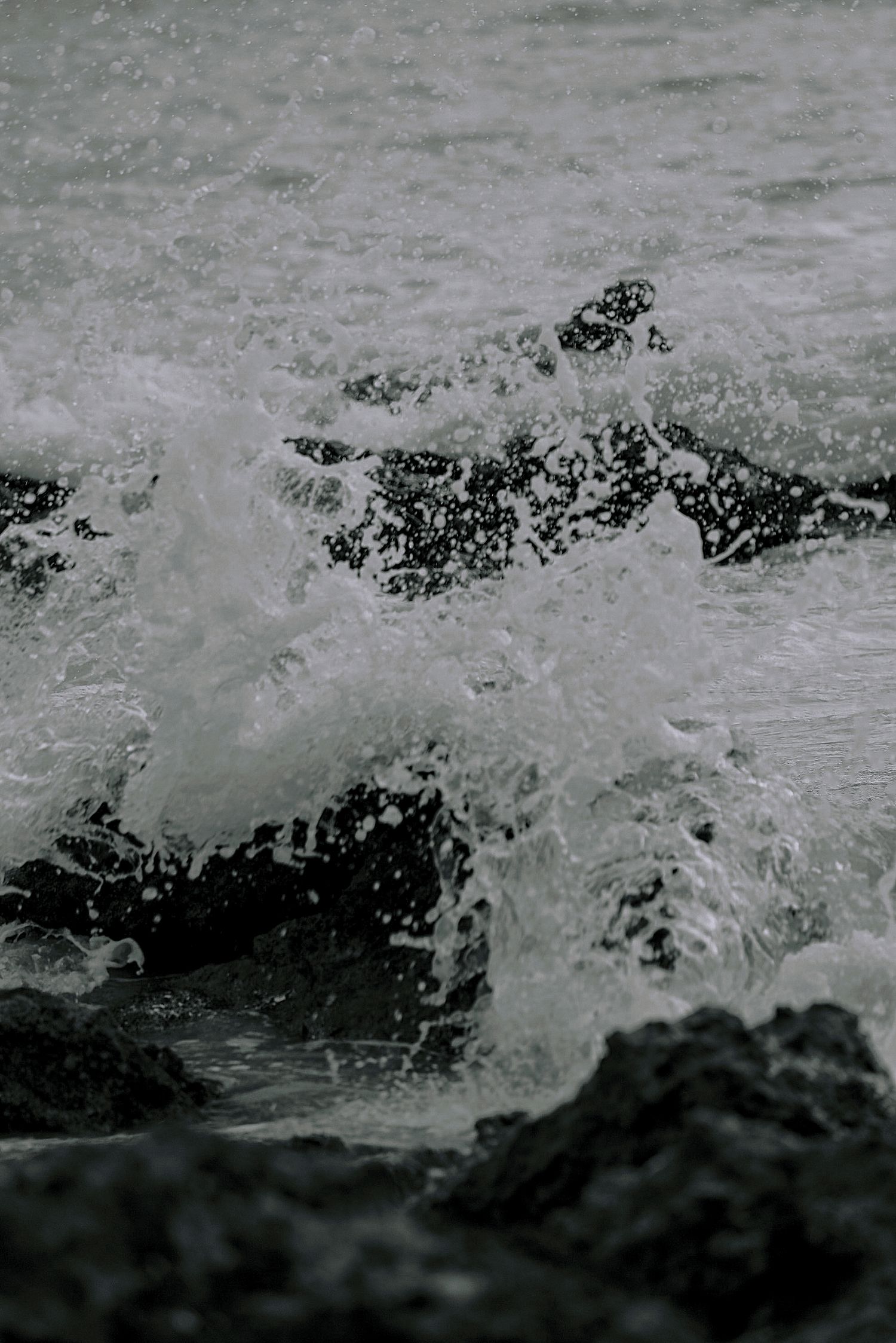 Ocean waves crashing dramatically against dark rocky shoreline, creating white sea foam and water spray in moody black and white tones.