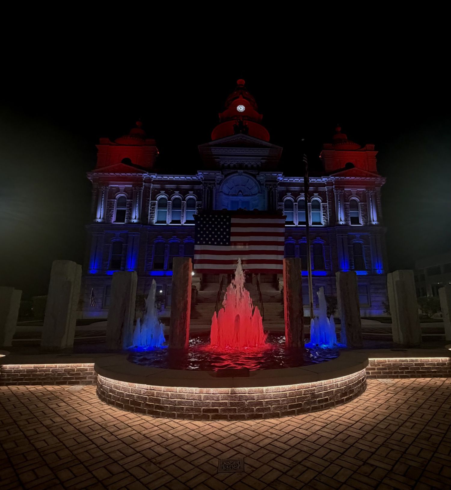 Troy, Ohio Historic courthouse lit in red, white, and blue at night with American flag and illuminated fountain display.