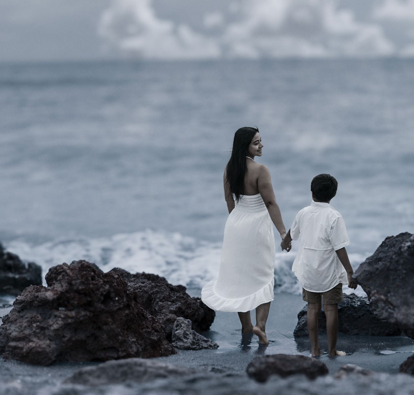 A woman in a white dress holds hands with a child near rocky shores of Caspersen Beach as ocean waves crash in the background.
