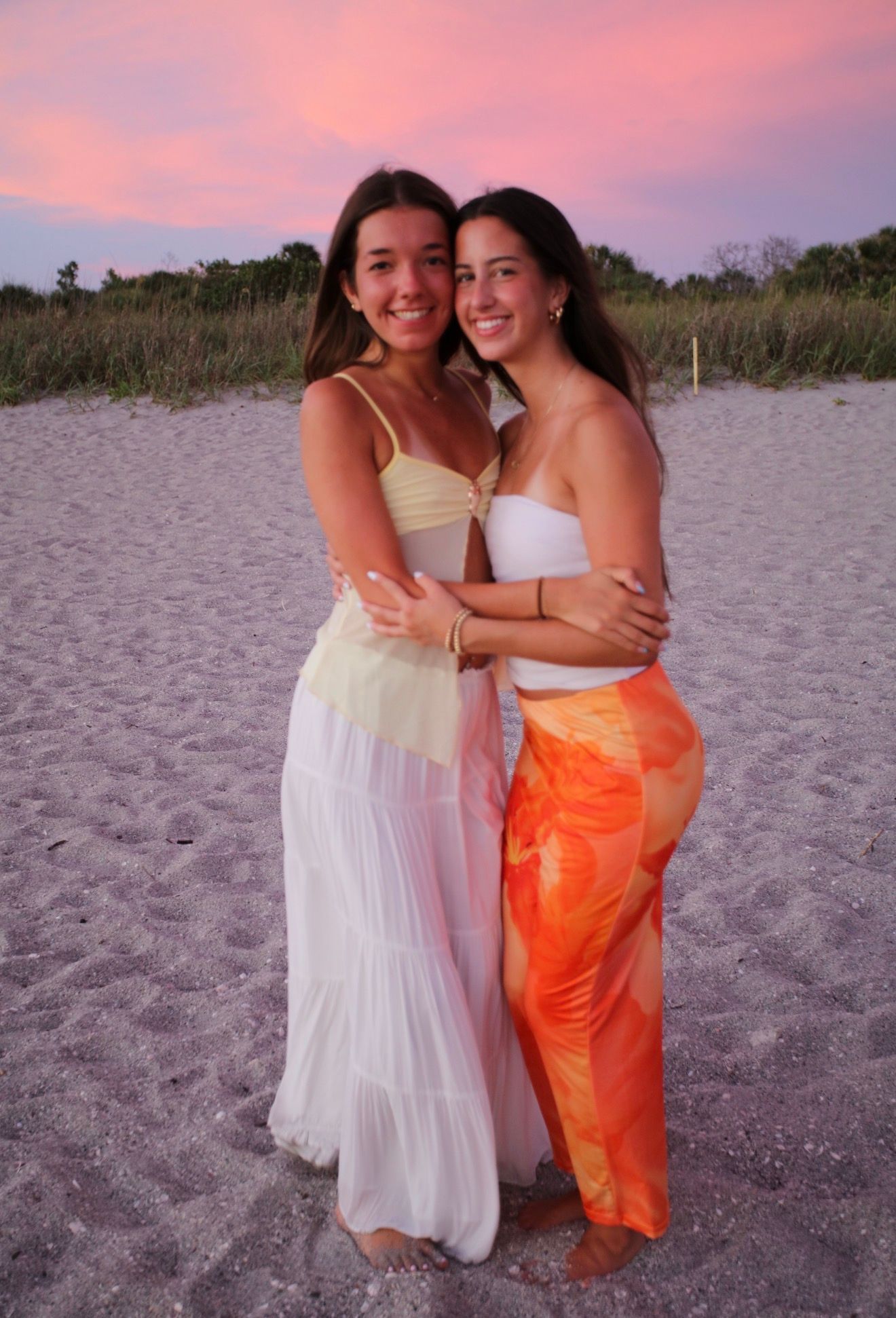 Two smiling young women hugging on sandy beach at pink sunset, wearing white and orange outfits.