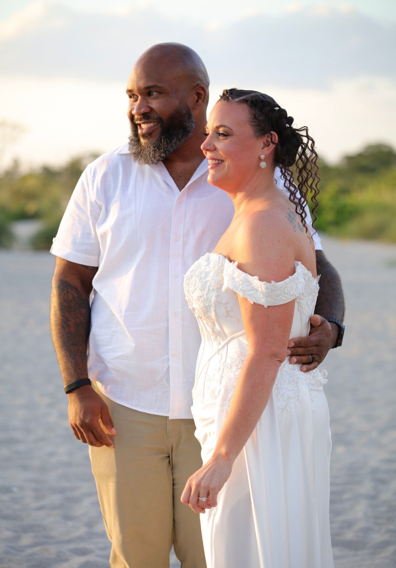 Couple in white wedding attire smiling on a sandy beach at sunset.