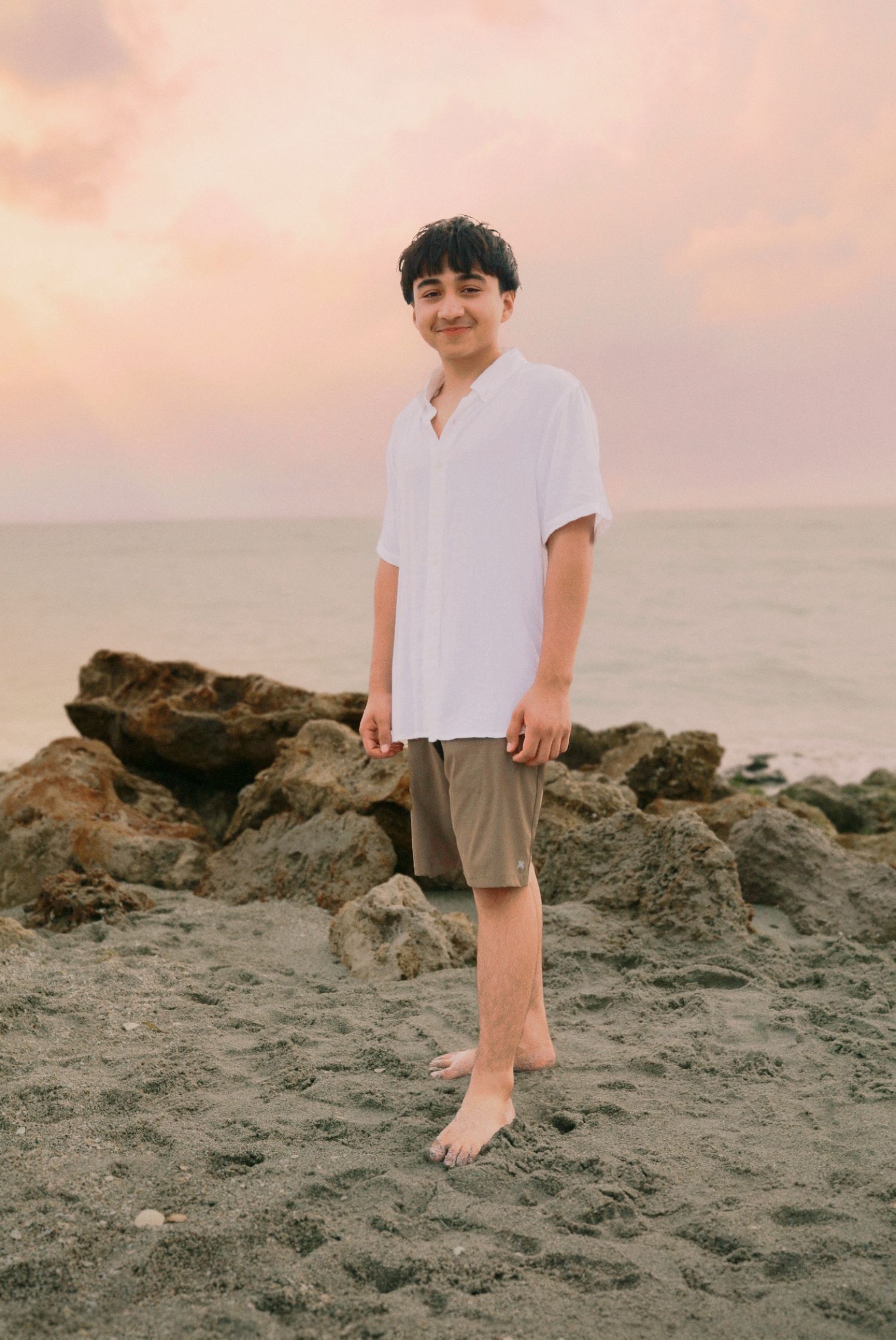 A smiling boy in a white shirt and khaki shorts stands barefoot on a sandy beach at sunset.