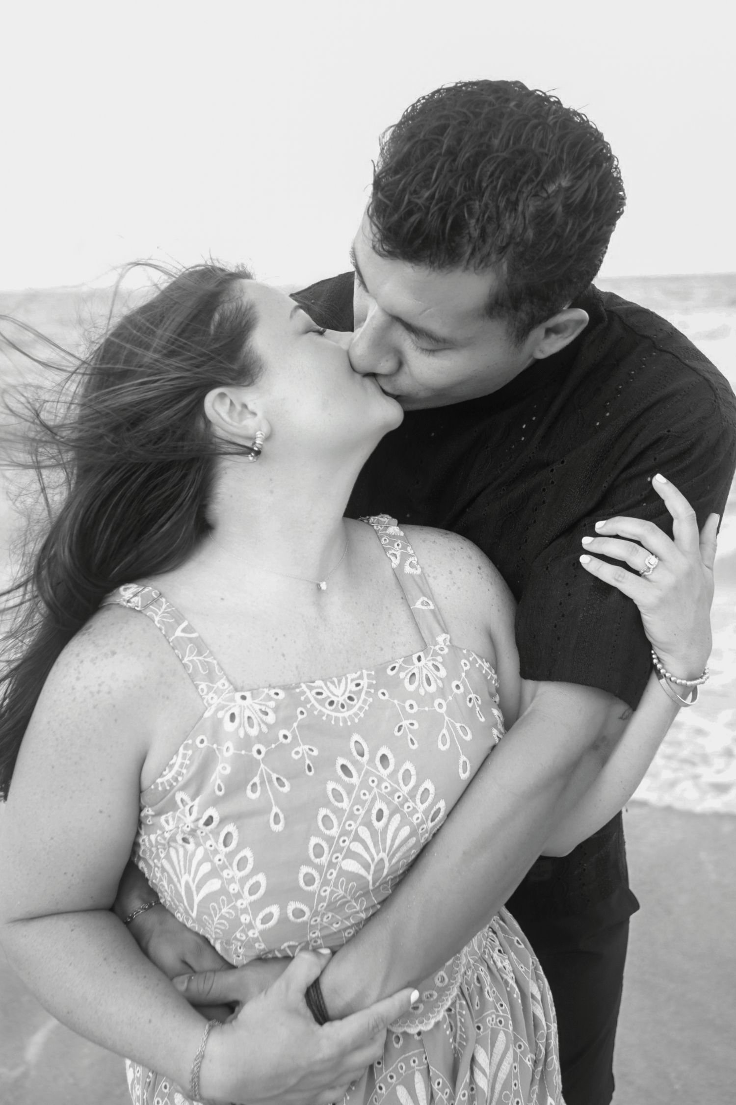 Black and white photo of couple kissing on beach, woman in floral dress with hair blowing in wind.