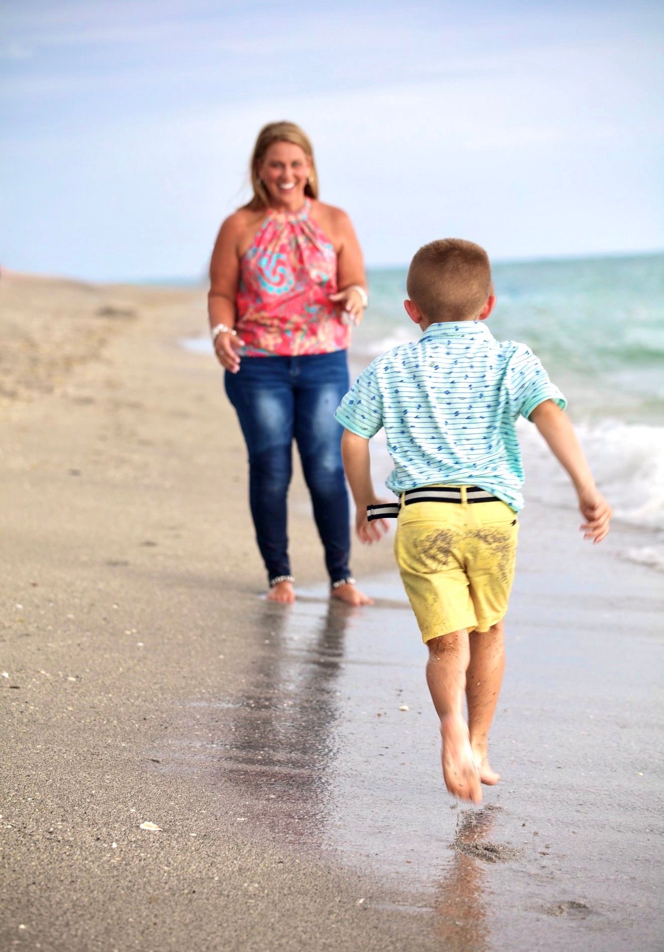 A young boy runs barefoot toward a smiling woman on a sunny beach, with ocean waves in the background.