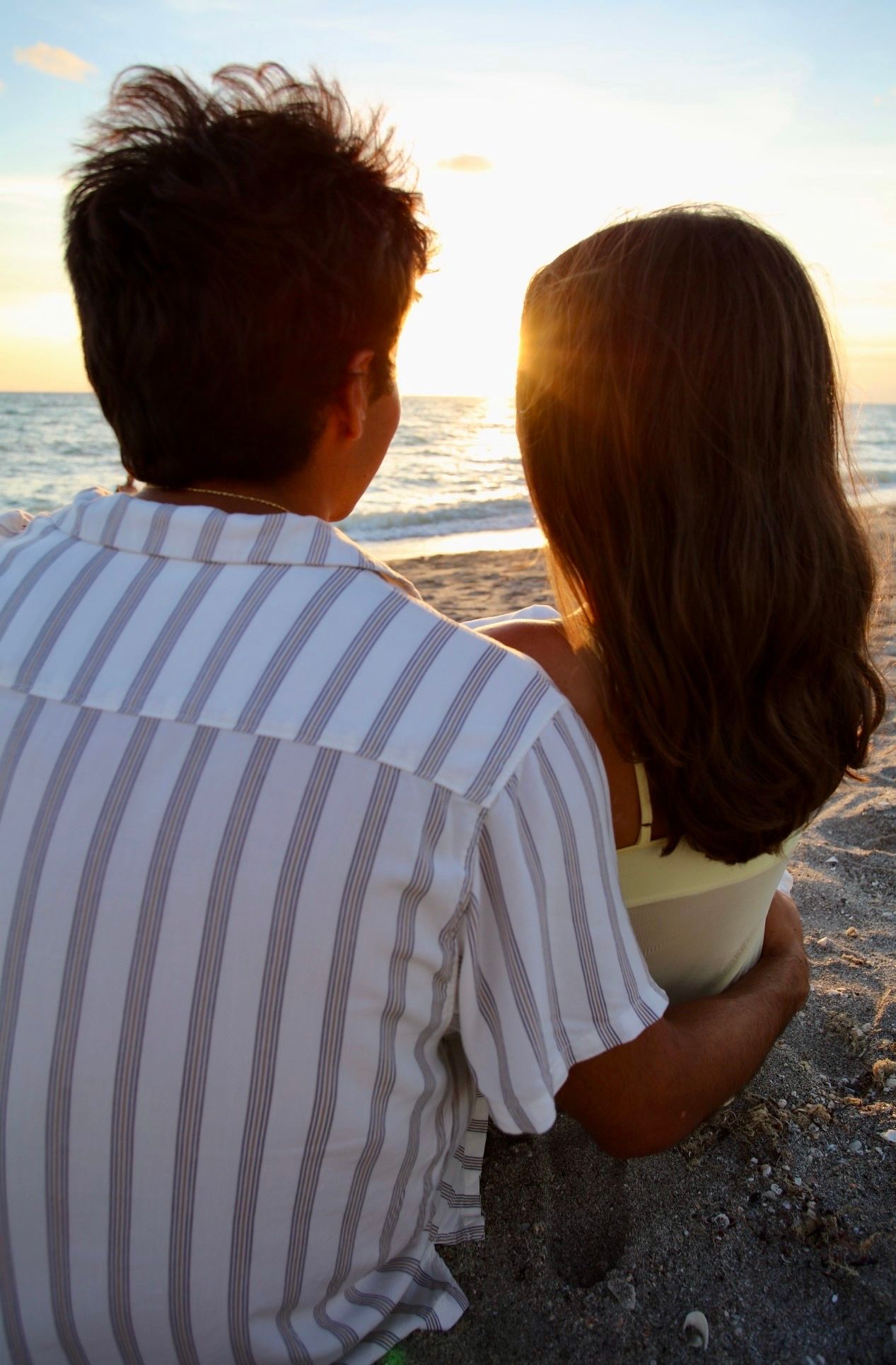 A couple sits together on a beach, watching the golden sunset over the calm ocean horizon.