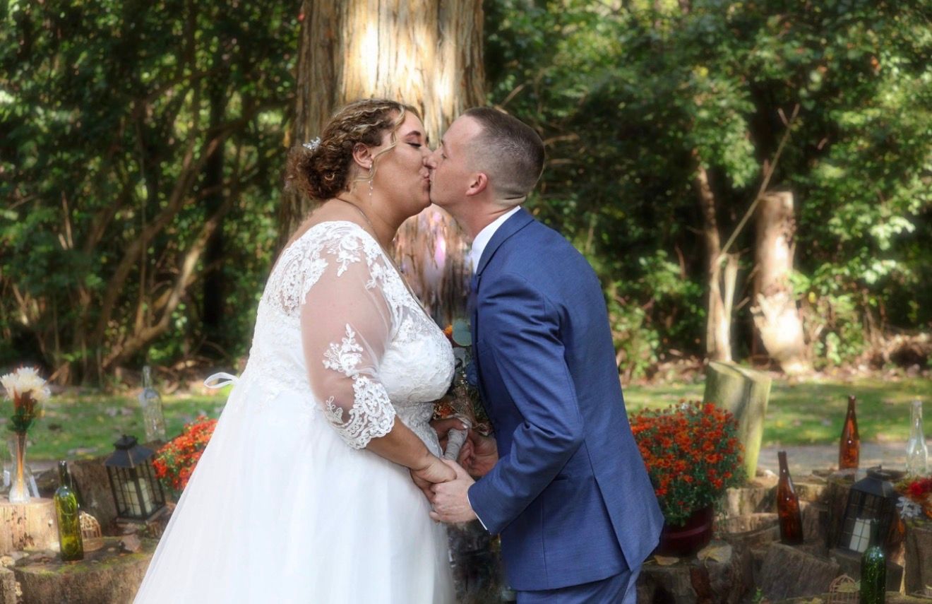 Bride and groom sharing a kiss at an outdoor wedding ceremony surrounded by lush green trees.