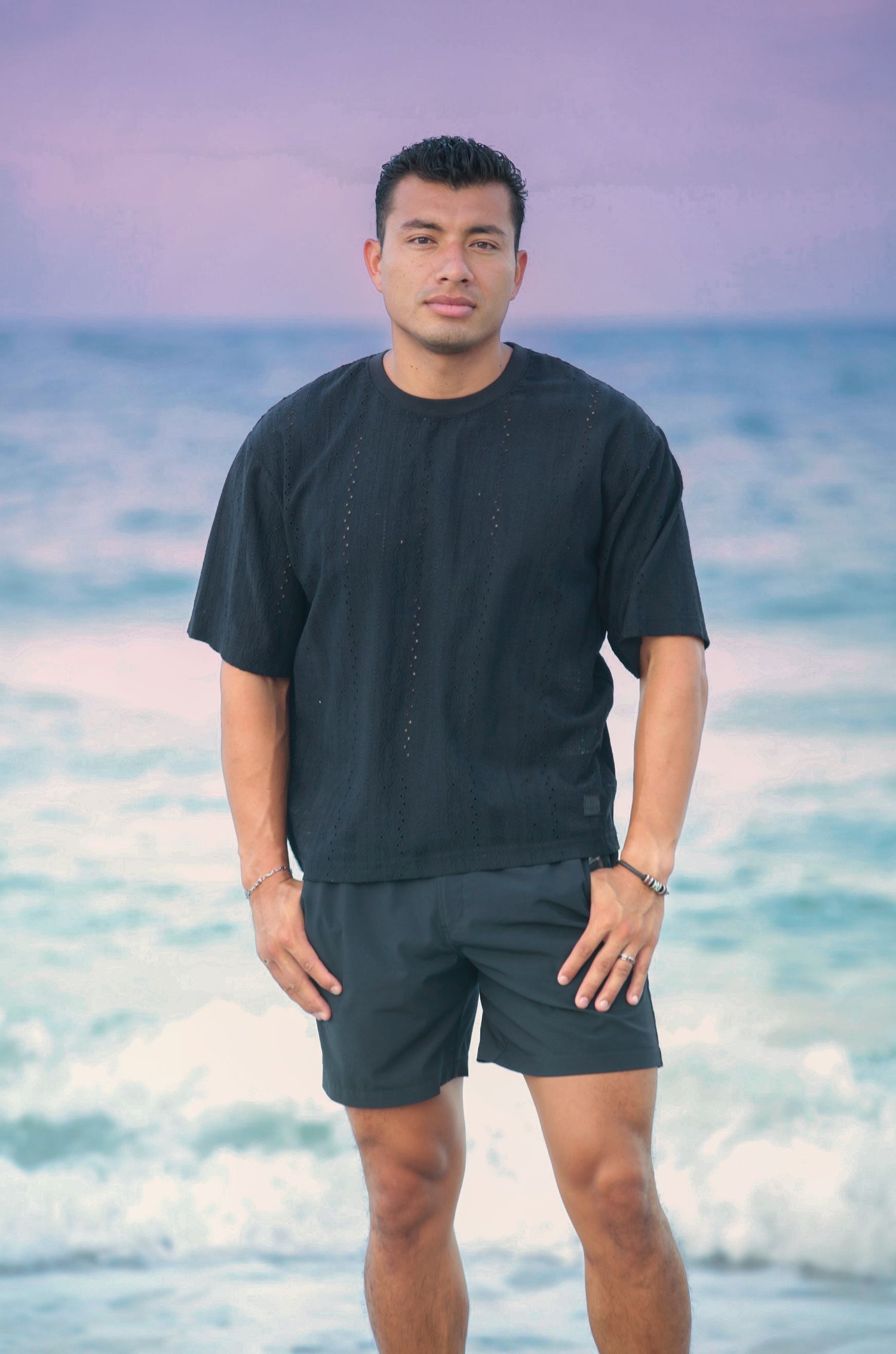 Man in black shirt and shorts standing on a beach with ocean waves and purple sky behind him.