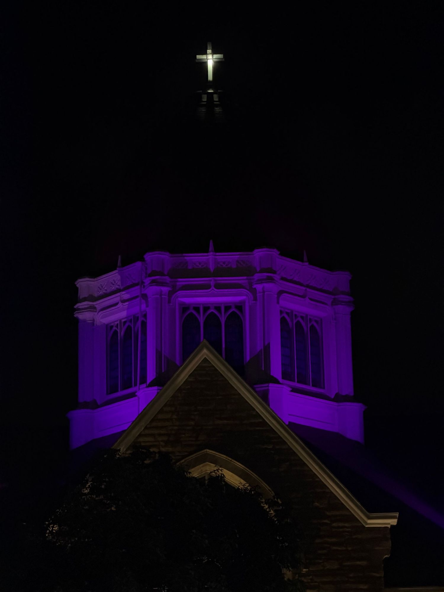 Church steeple dramatically illuminated in purple light at night with glowing white cross on top.