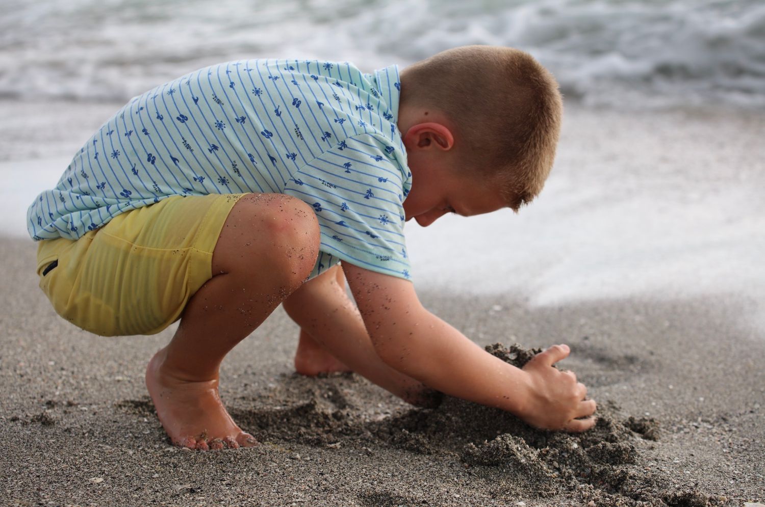A young boy crouches on a sandy beach, playing and digging in the wet sand near the ocean shoreline.