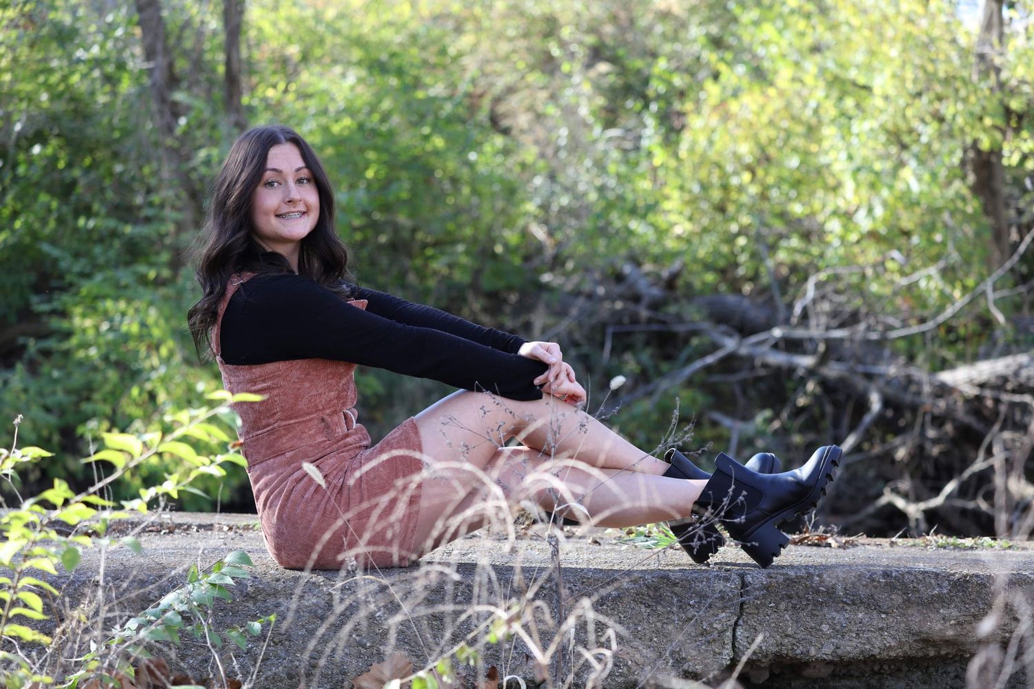 Young woman in rust dress and black boots sits on stone wall surrounded by lush green forest.