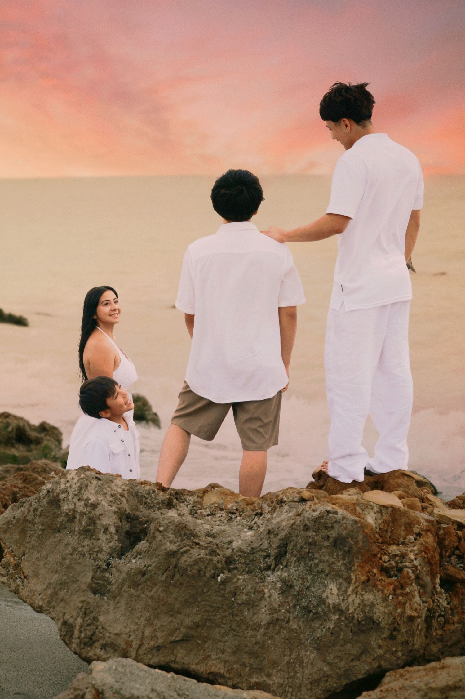 Family of four standing on rocks at a beach during a colorful sunset, dressed in white.