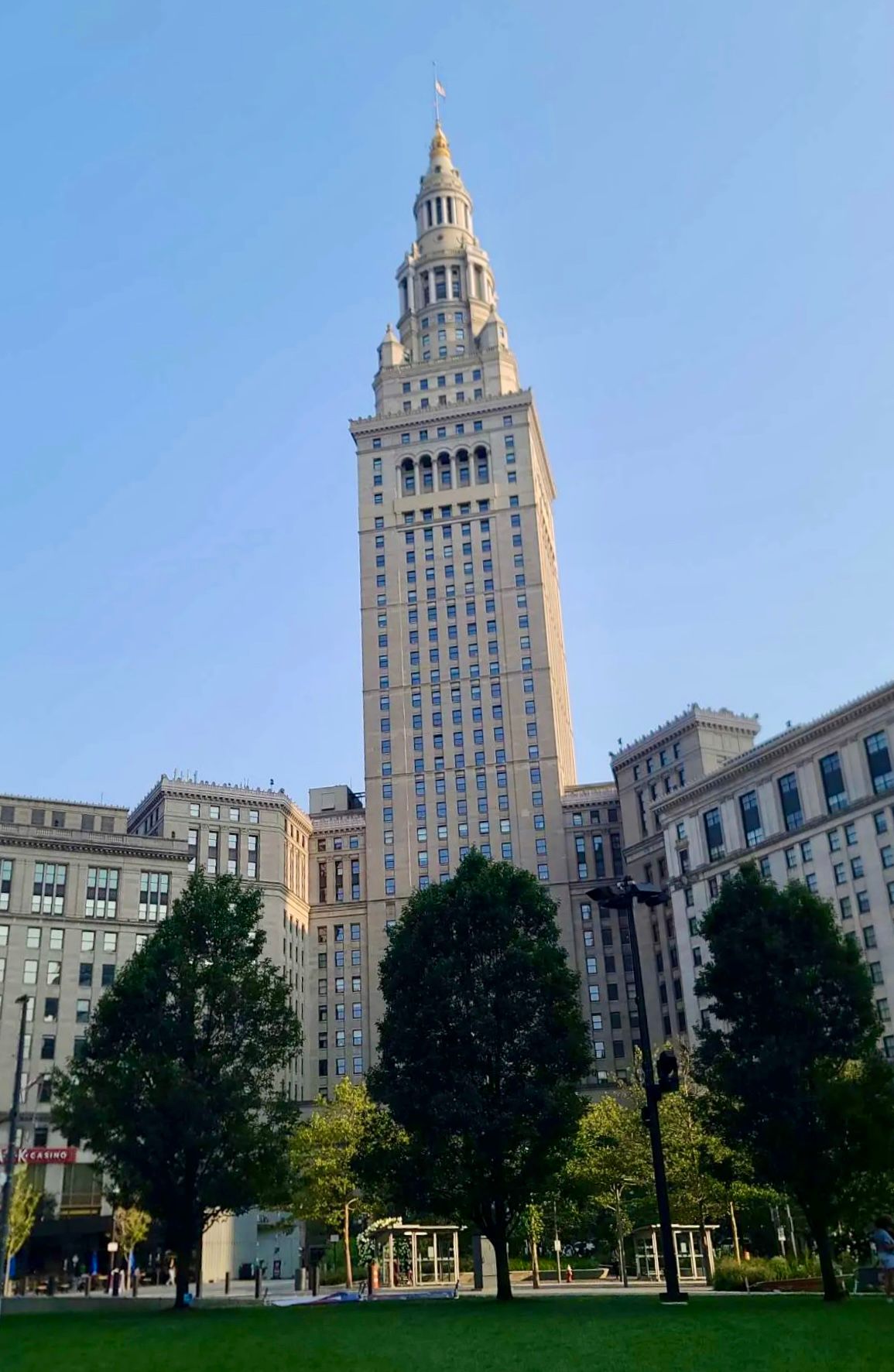 Terminal Tower skyscraper rises above green trees in Cleveland, Ohio, against a clear blue sky.
