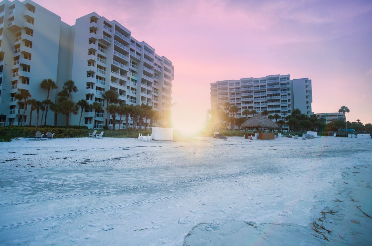 Beachfront hotels glow under a pastel pink and purple sunset sky on a white sandy Florida beach.