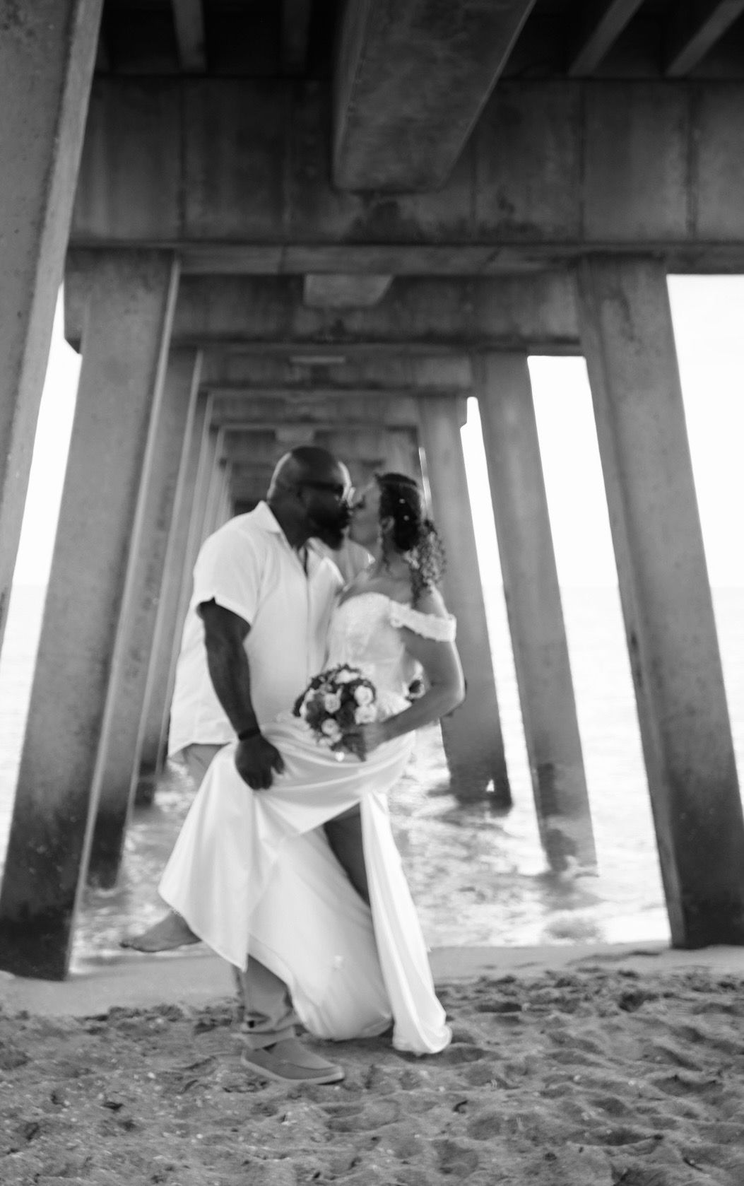 Black and white photo of a couple kissing in wedding attire under a beach pier, bride holding a bouquet.