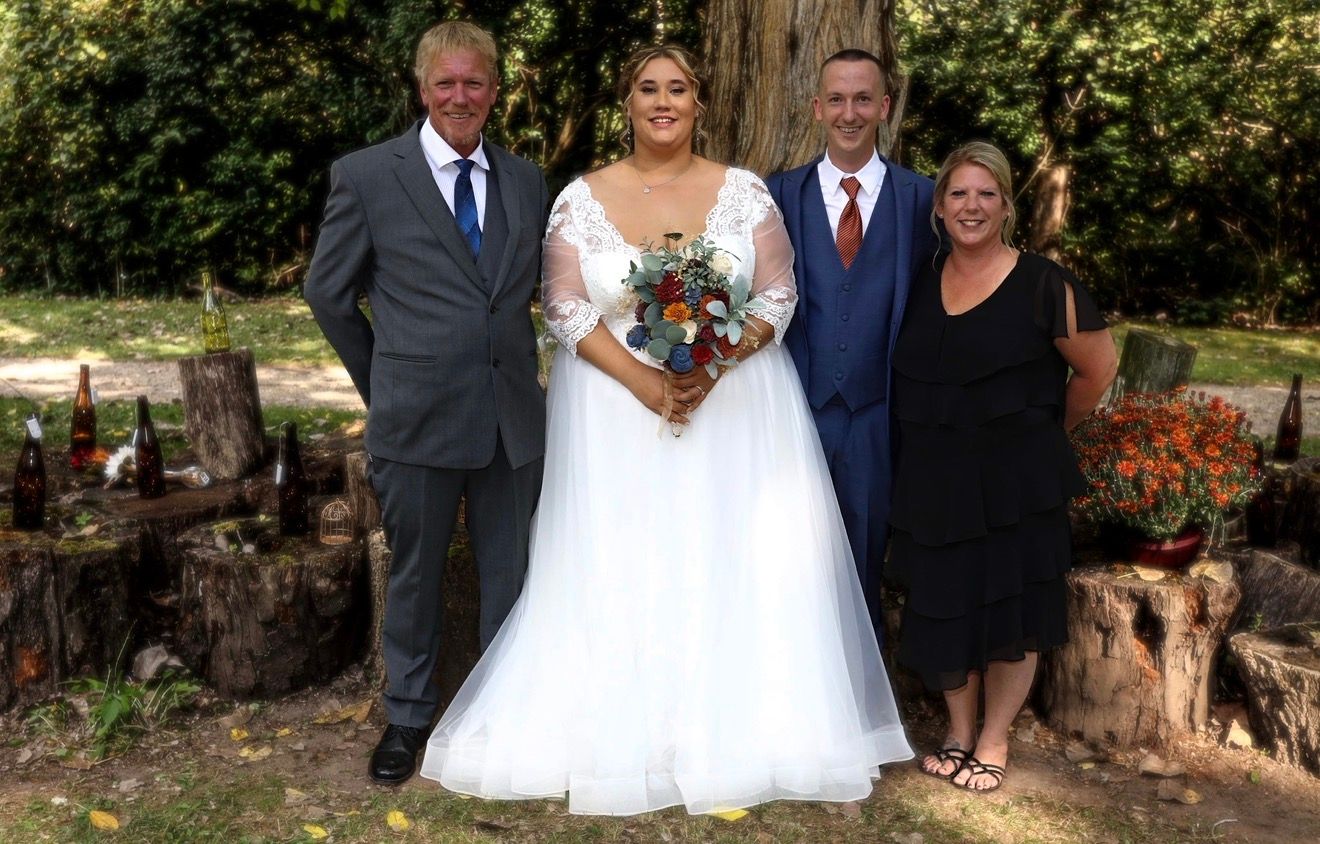 Bride in white tulle gown poses with three guests holding a colorful bouquet at an outdoor woodland wedding.