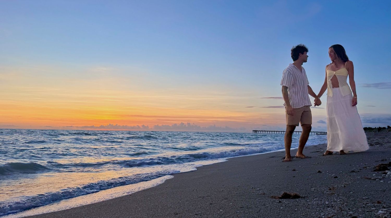 Couple holds hands walking along shoreline at golden sunset on a sandy beach.