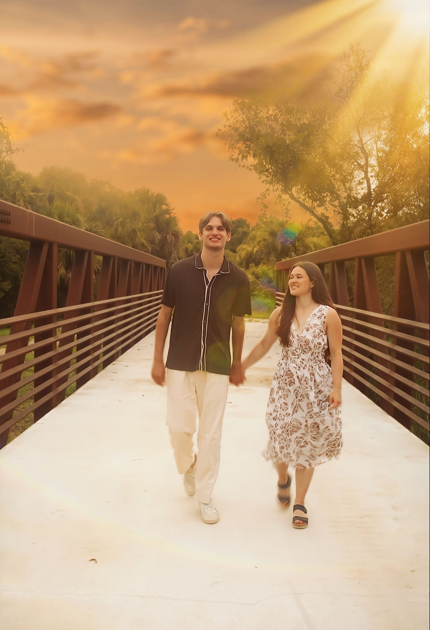 A couple walks hand-in-hand on a bridge at sunset, surrounded by lush trees and golden sunlight.
