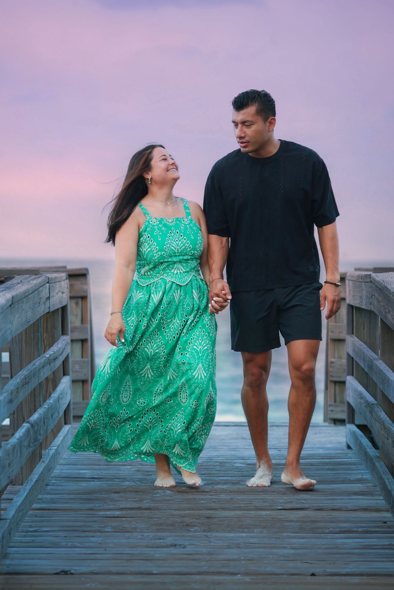 Couple holding hands walking on wooden pier at sunset, woman in green dress.