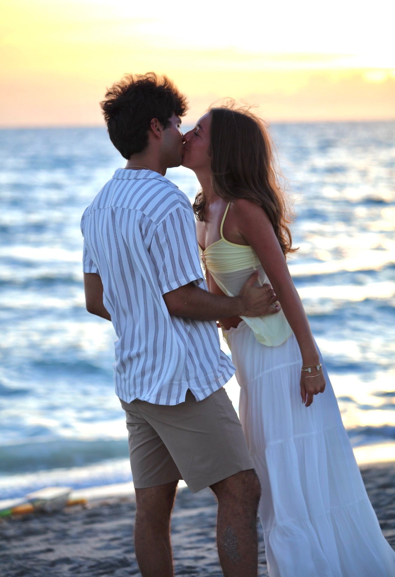 A romantic young couple shares a kiss on the beach at sunset, the woman in a yellow top and white skirt.