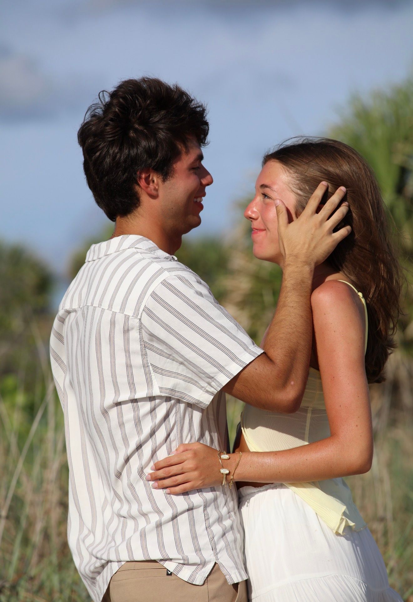 Young romantic couple embracing outdoors near beach dunes, man tenderly holding woman's face in golden light.
