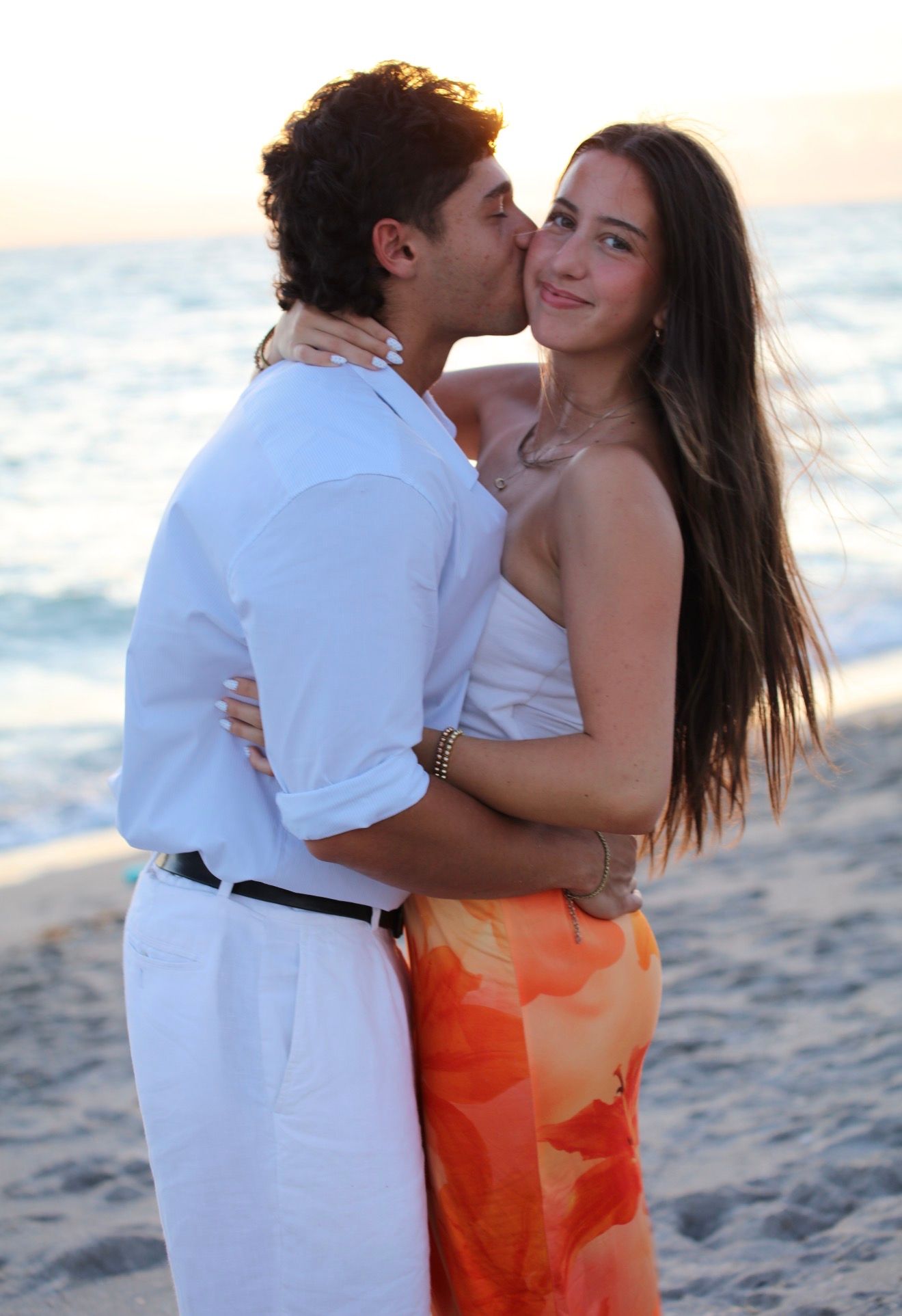 Young man kissing smiling woman on cheek at beach sunset, she wears orange floral pants.