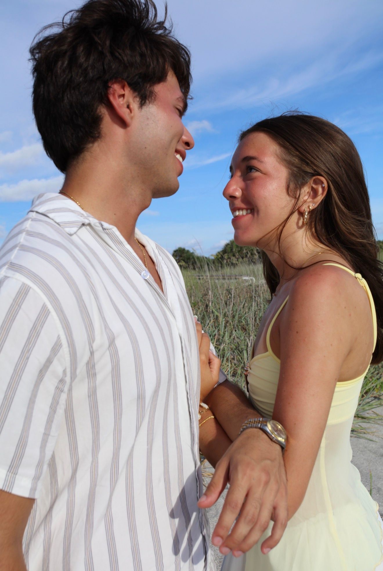 A smiling young couple gazes at each other outdoors on a sunny beach day, woman in yellow dress, man in striped shirt.