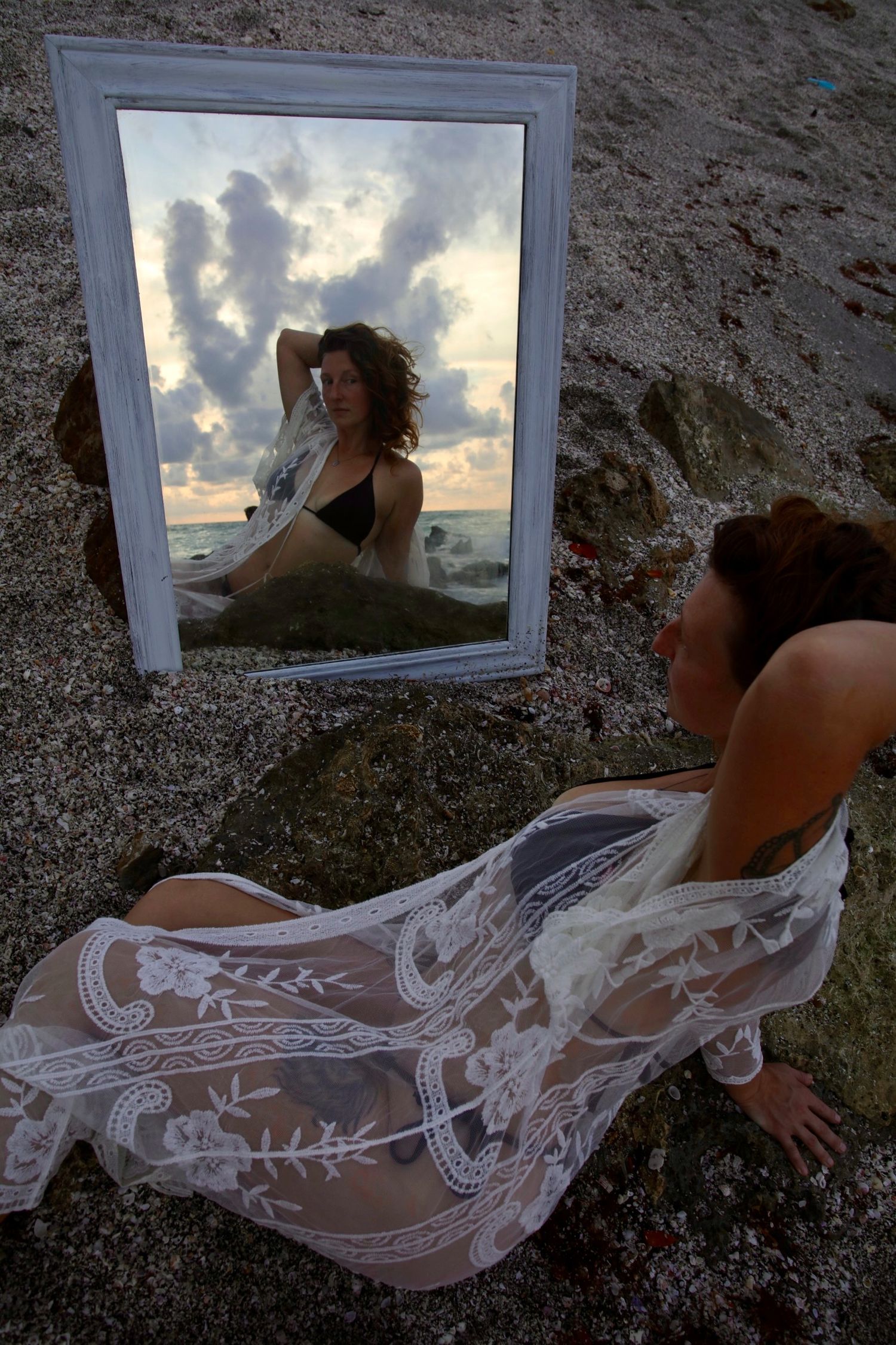 Woman in lace cover-up gazes into mirror on rocky beach reflecting sunset sky.