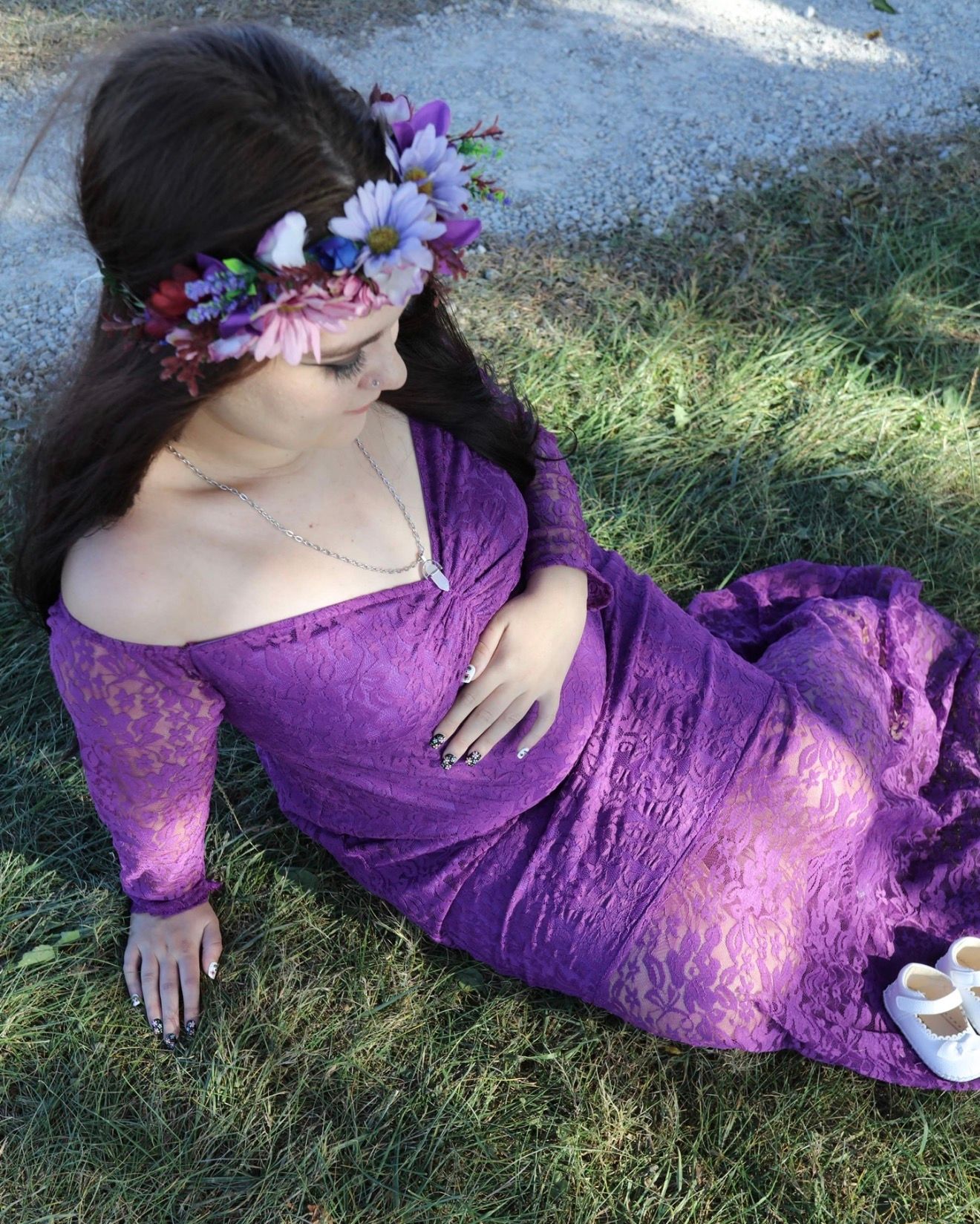 Pregnant woman in purple lace off-shoulder gown with floral crown sits gracefully on green grass.