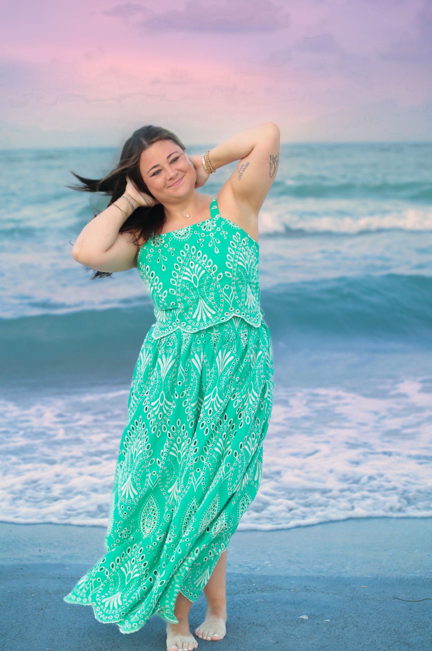 Woman in green patterned maxi dress posing on beach at sunset with ocean waves in background.