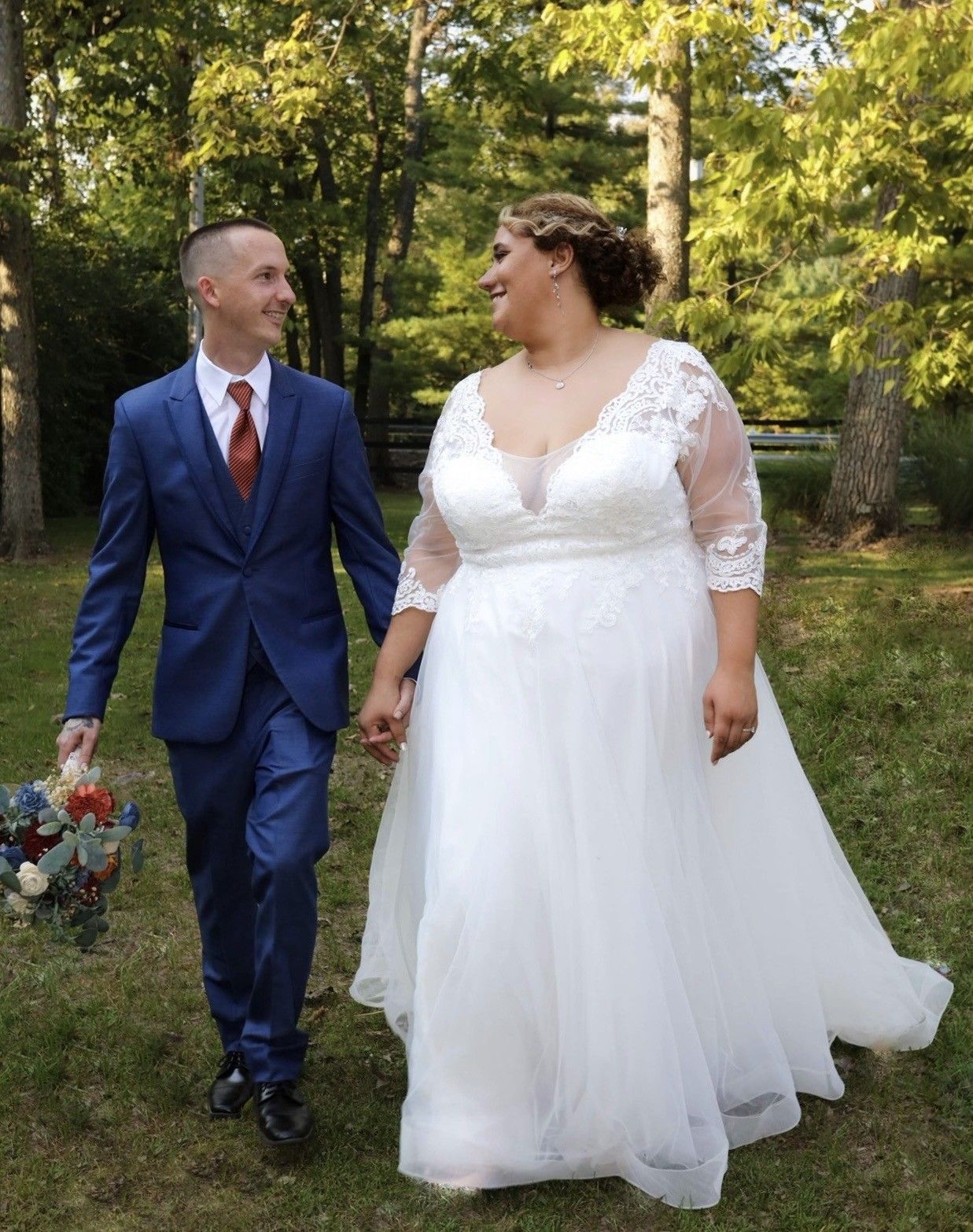 Bride and groom walking hand in hand outdoors, she in a lace wedding gown, he in a blue suit with red tie.