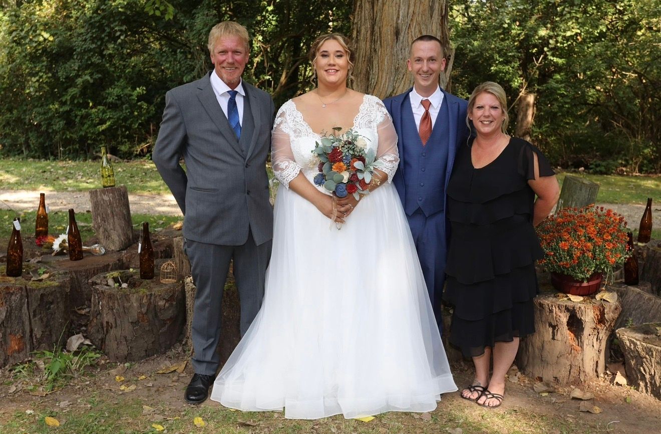 Wedding party of four posing outdoors with bride in white gown holding colorful bouquet, surrounded by rustic tree stump decor.