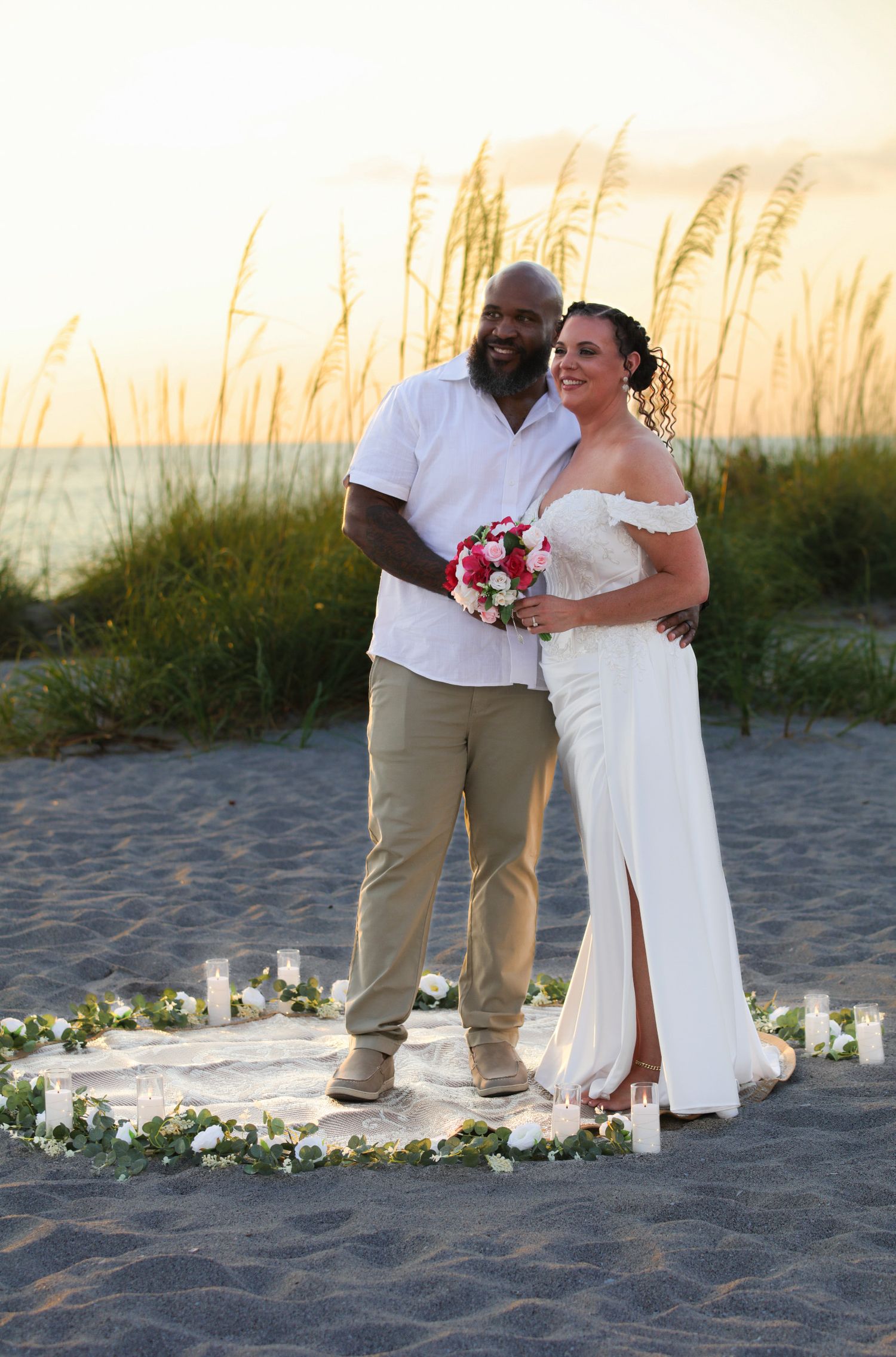 Couple poses on sandy beach at sunset, bride in white gown holding pink bouquet, surrounded by candles and floral arrangement.