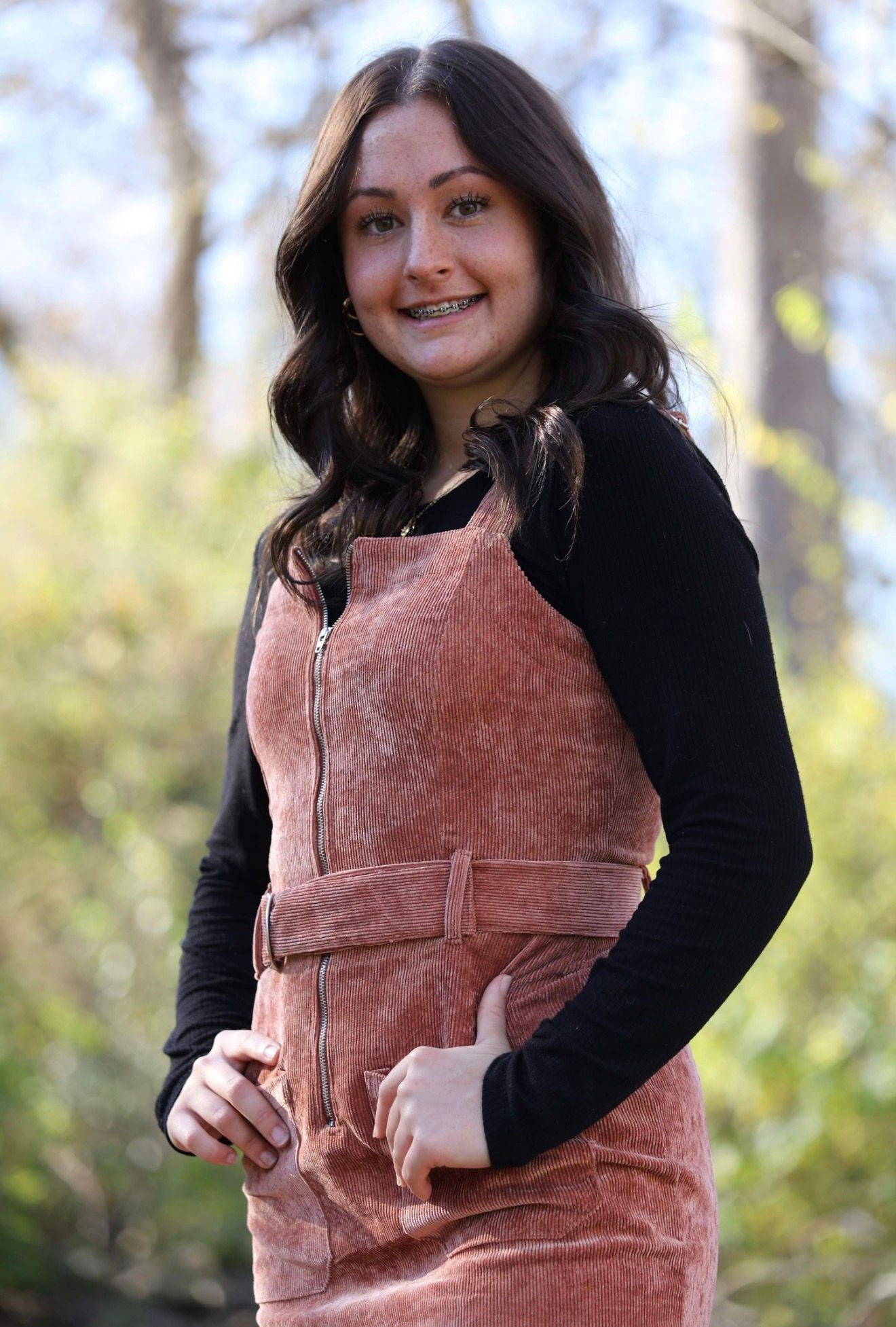 Smiling teenage girl with braces wearing a pink velvet overall dress over black long-sleeve shirt outdoors.