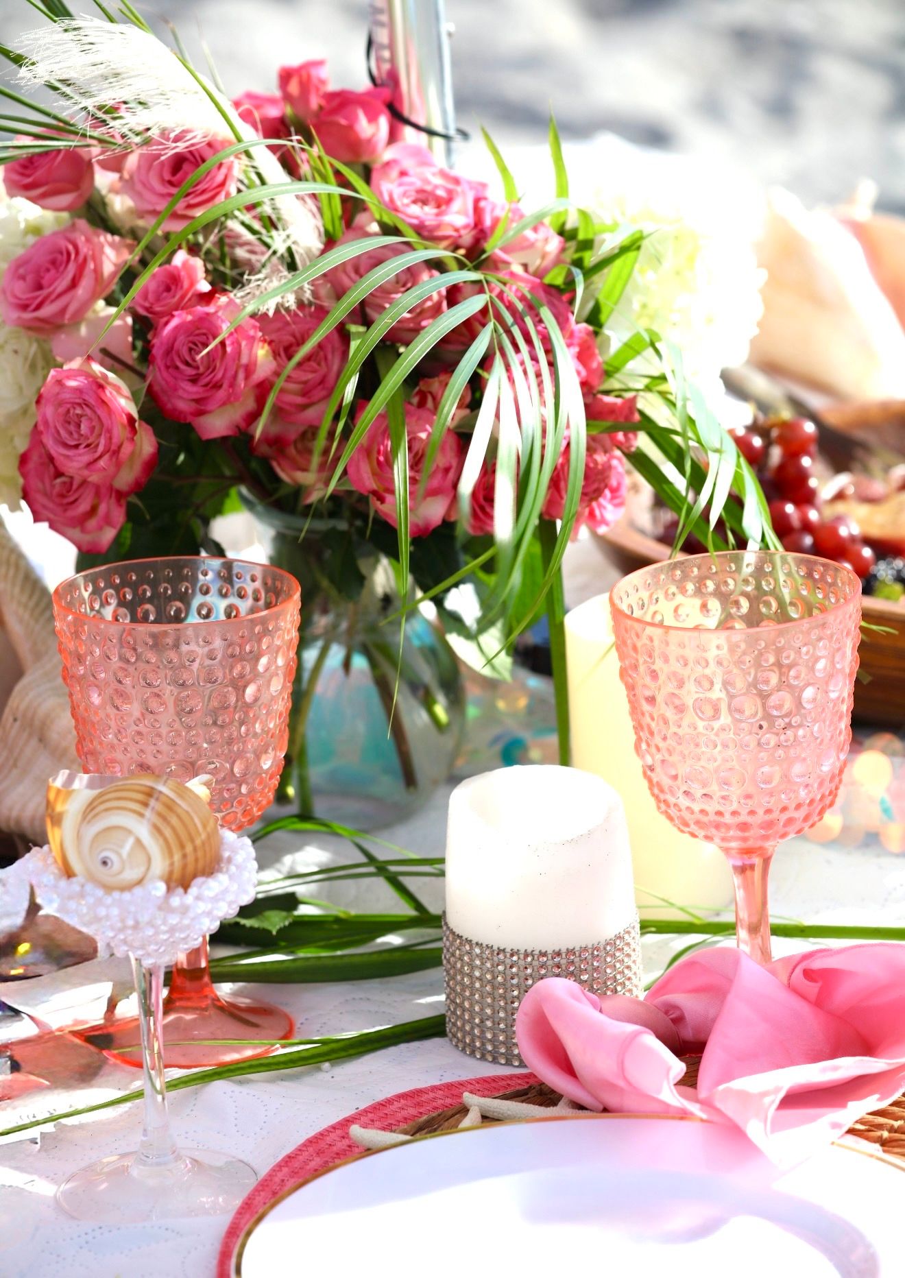 Pink hobnail goblets and roses centerpiece on elegant beach table setting with candles and seashells.